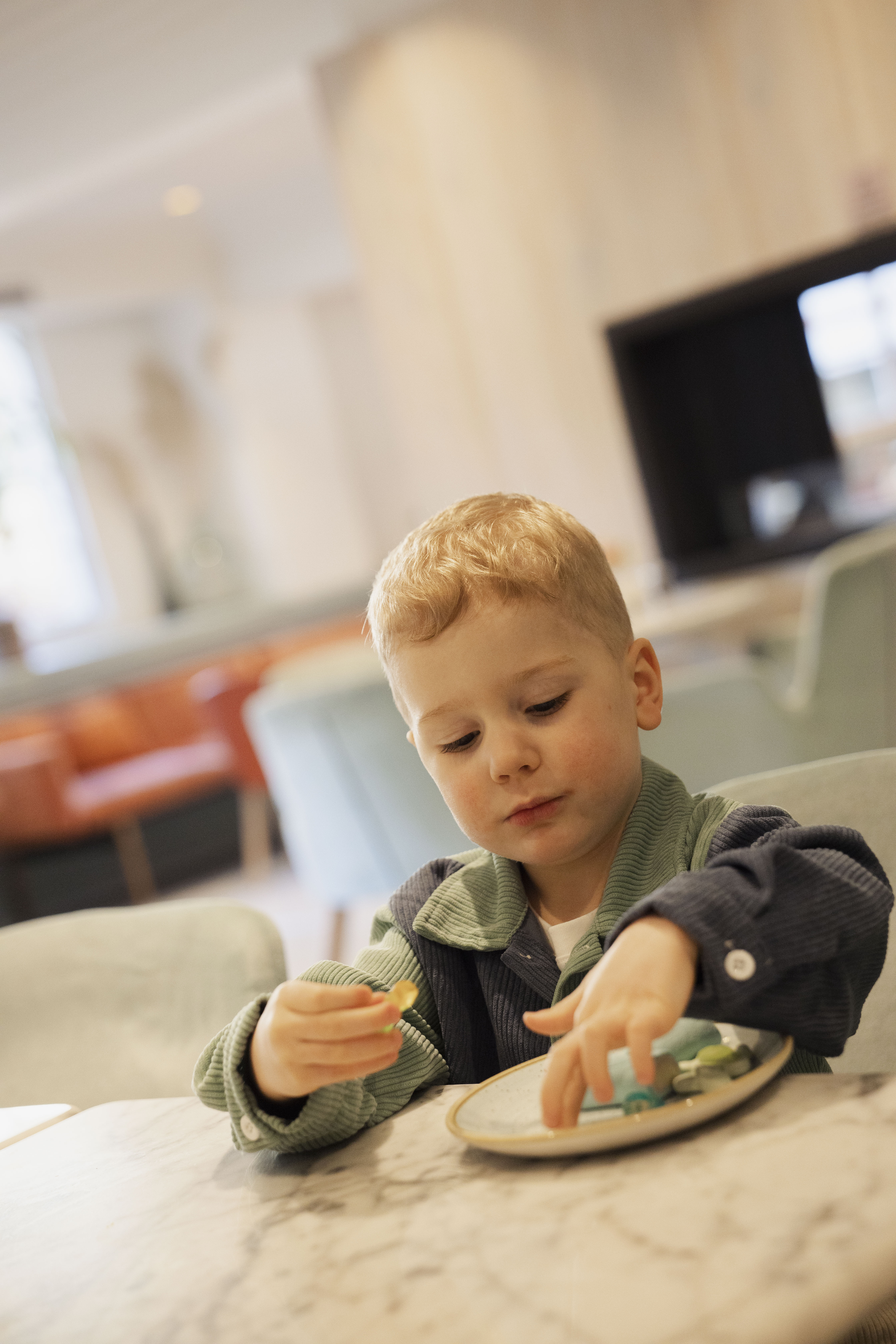Young blonde boy playing with candies on a plate at a cafe table.