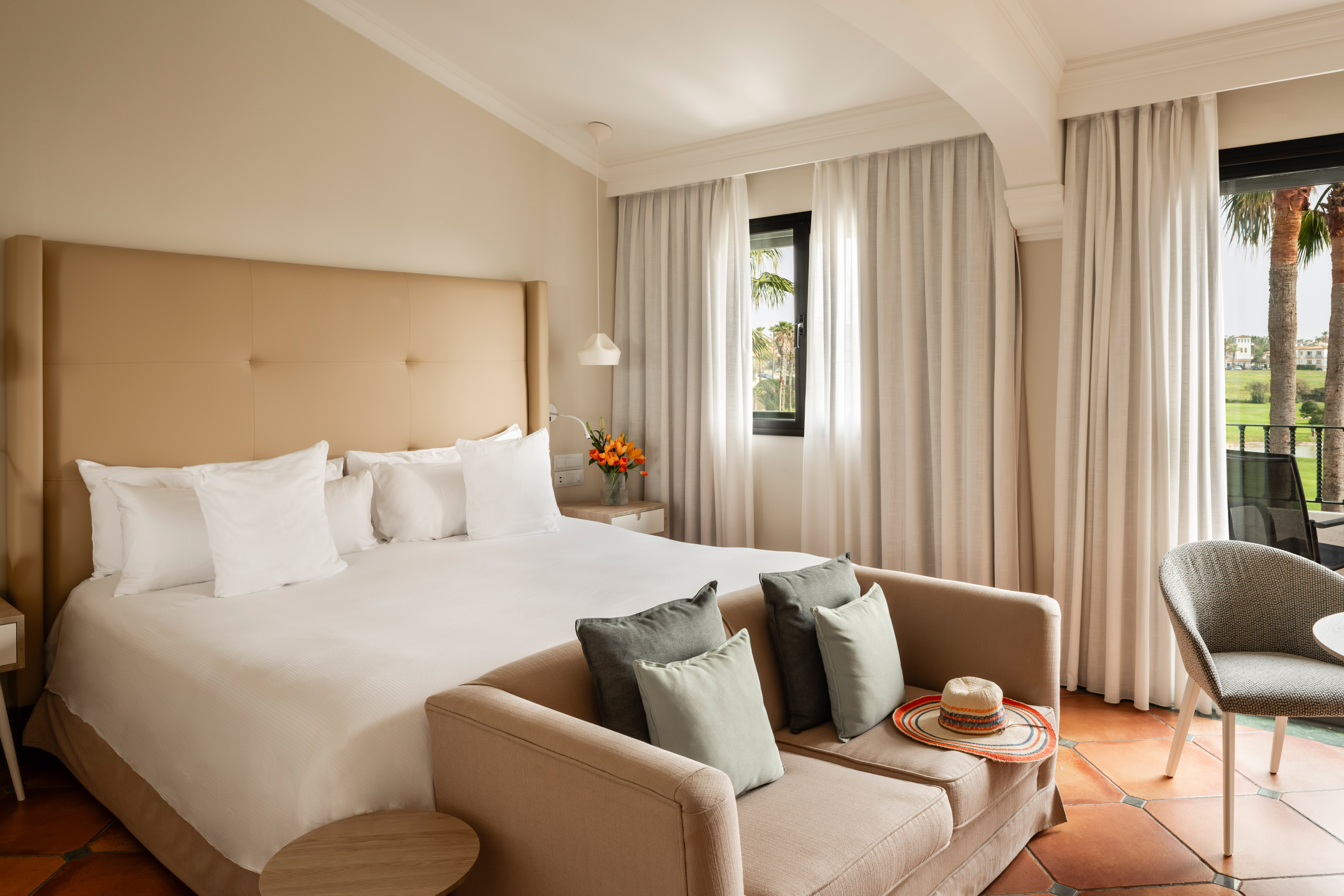 Elegant hotel room with king bed, sofa, and balcony overlooking tropical resort.