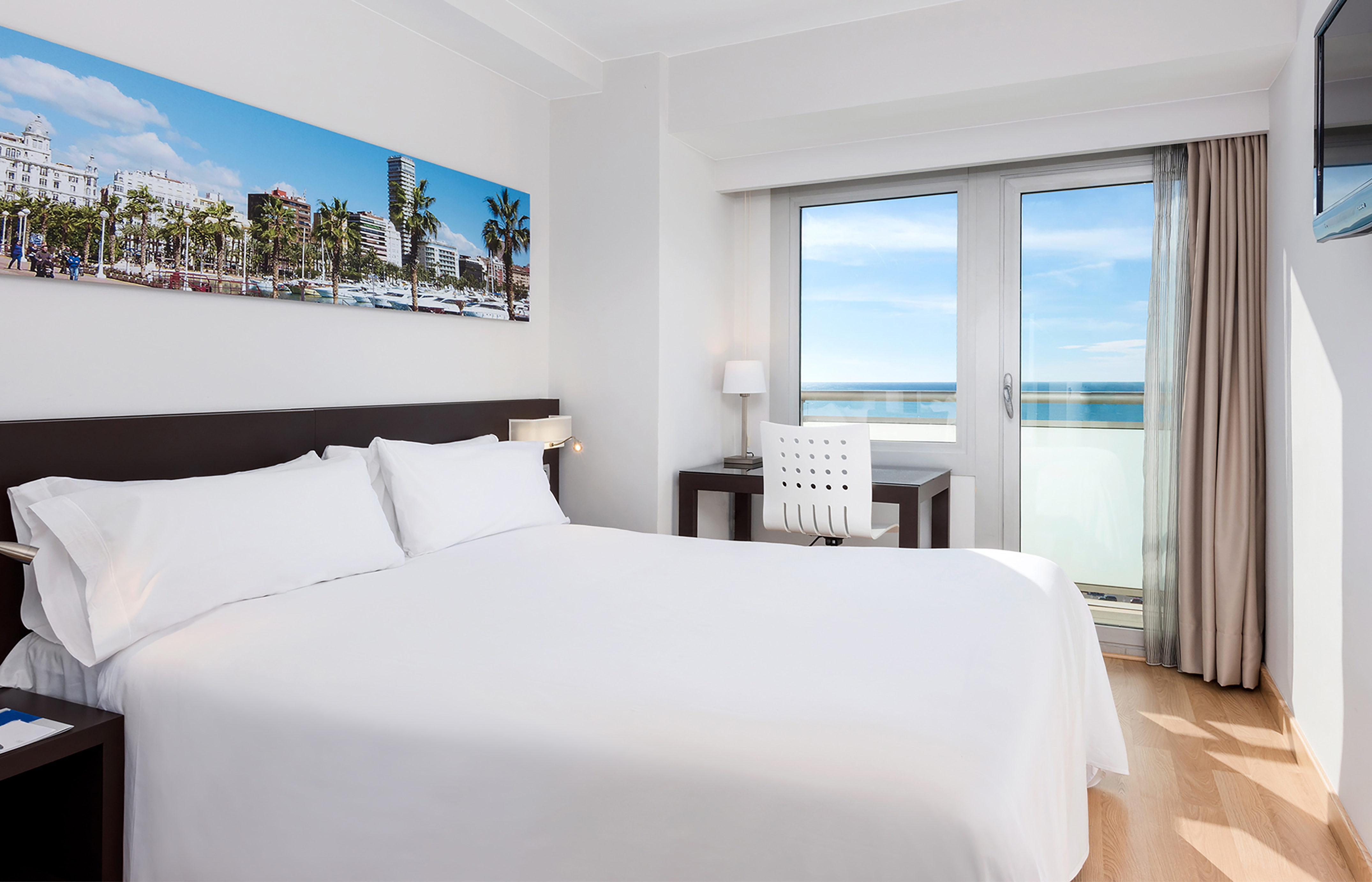 Modern hotel room with ocean view balcony, king bed, desk, and city artwork.