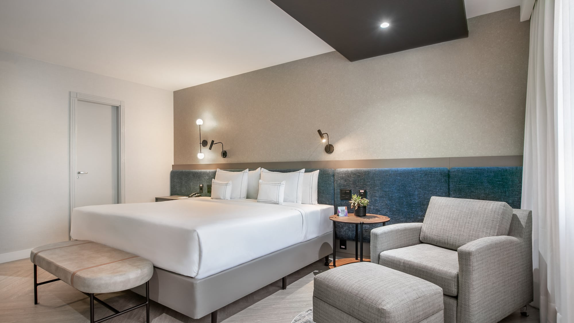 Modern hotel bedroom with king bed, blue headboard, grey armchair, and ottoman.