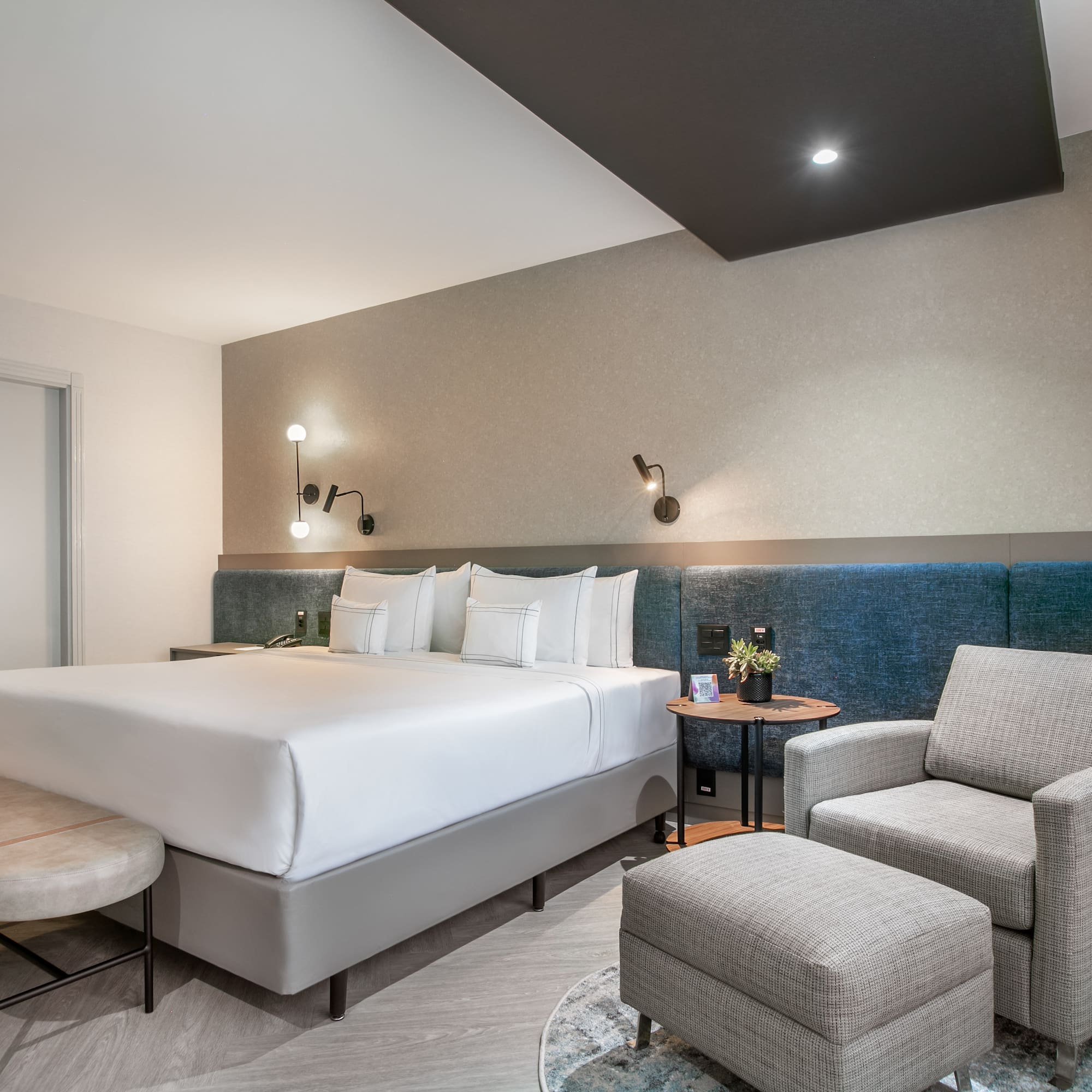 Modern hotel bedroom with king bed, blue headboard, armchair, and sleek design.
