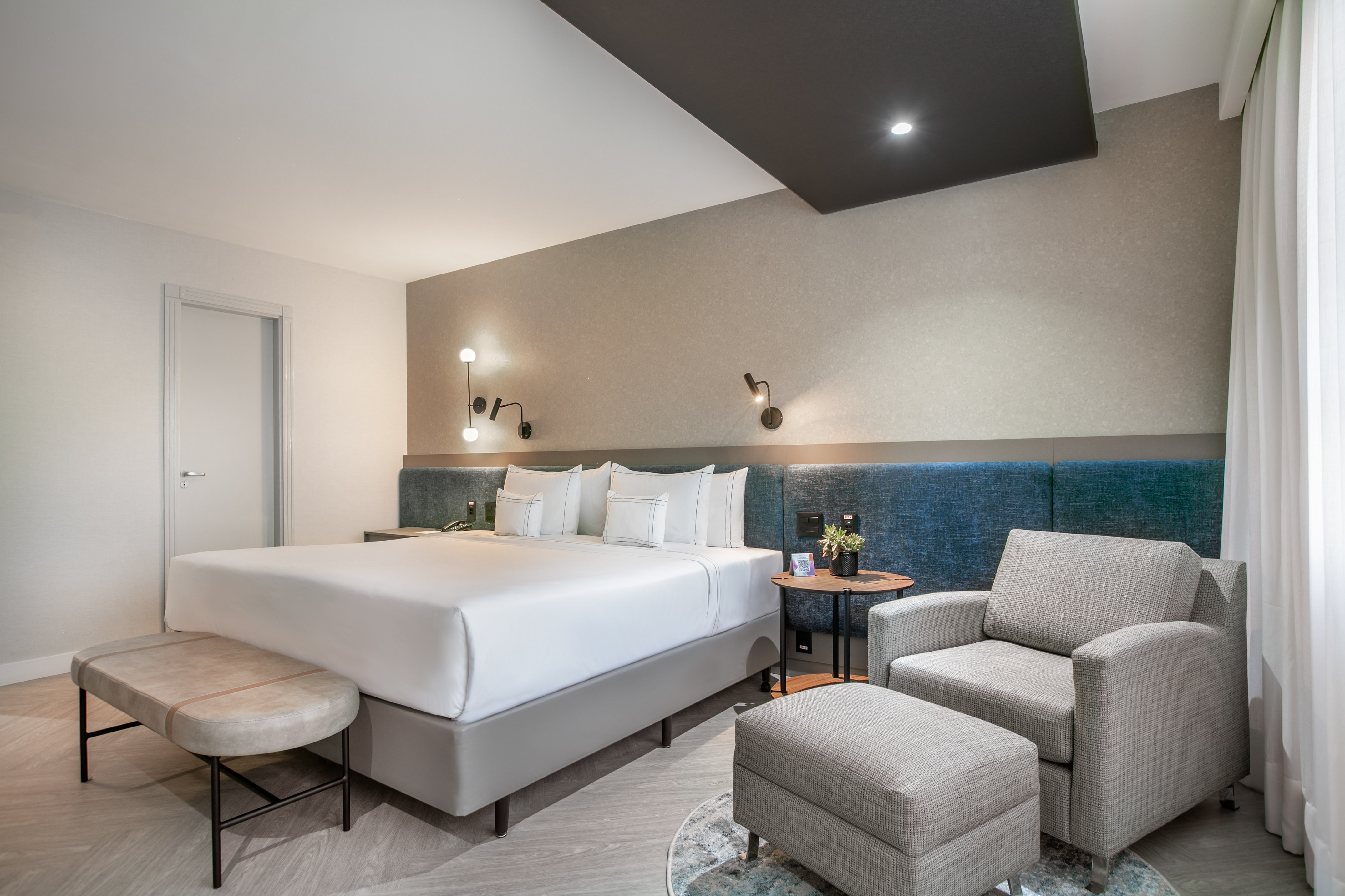 Modern hotel bedroom with king bed, blue headboard, armchair, and sleek design.
