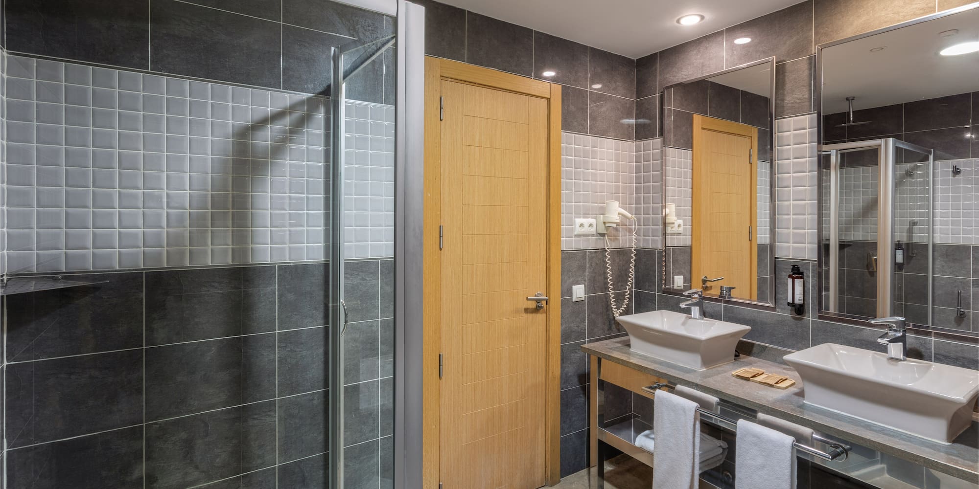 Modern hotel bathroom with gray tiles, glass shower, and double sink vanity.