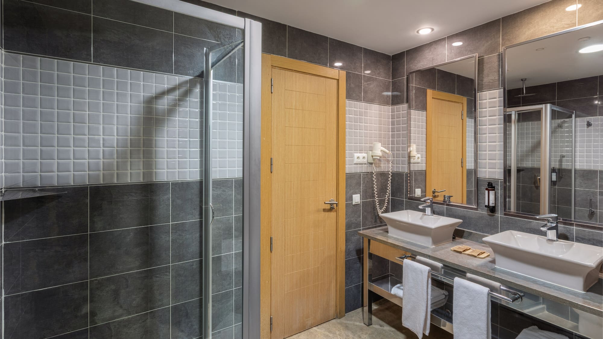 Modern hotel bathroom with gray tiles, glass shower, and double sink vanity.