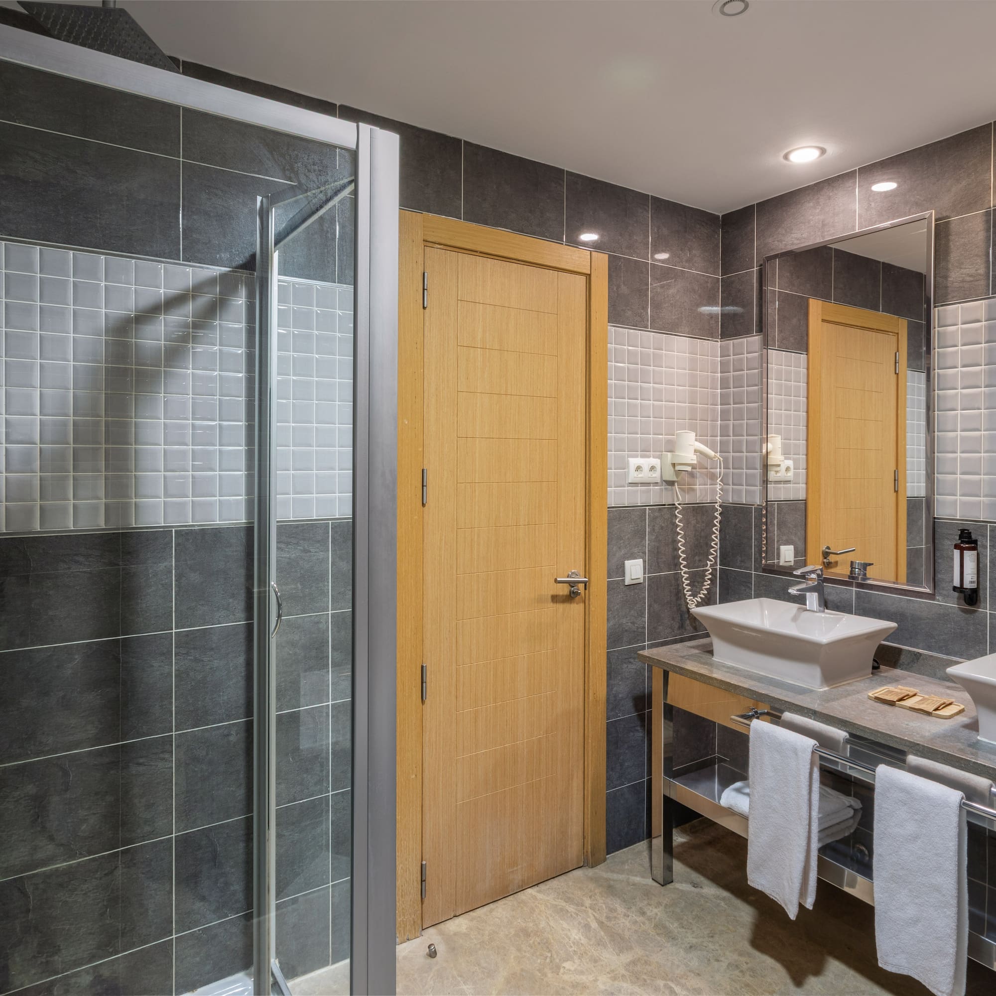 Modern hotel bathroom with gray tiles, glass shower, and double sink vanity.