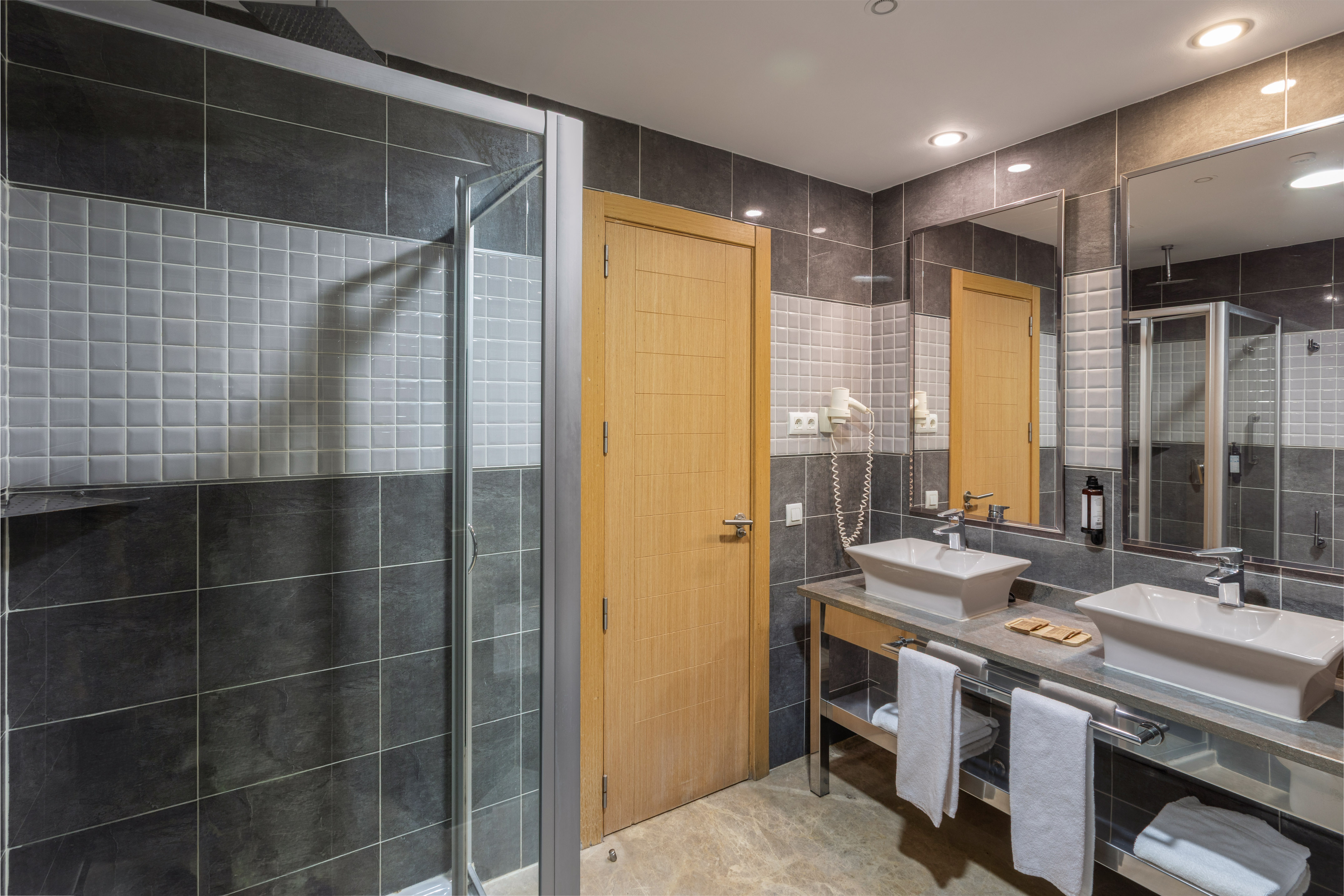 Modern hotel bathroom with gray tiles, glass shower, double vanity, vessel sinks.