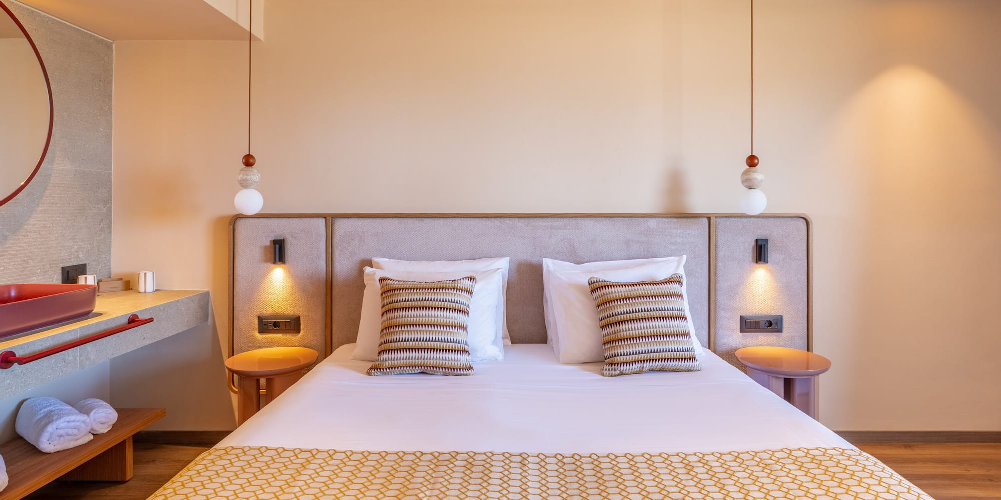 Modern hotel bedroom with patterned bed, pendant lights, and modern red-basin ensuite.