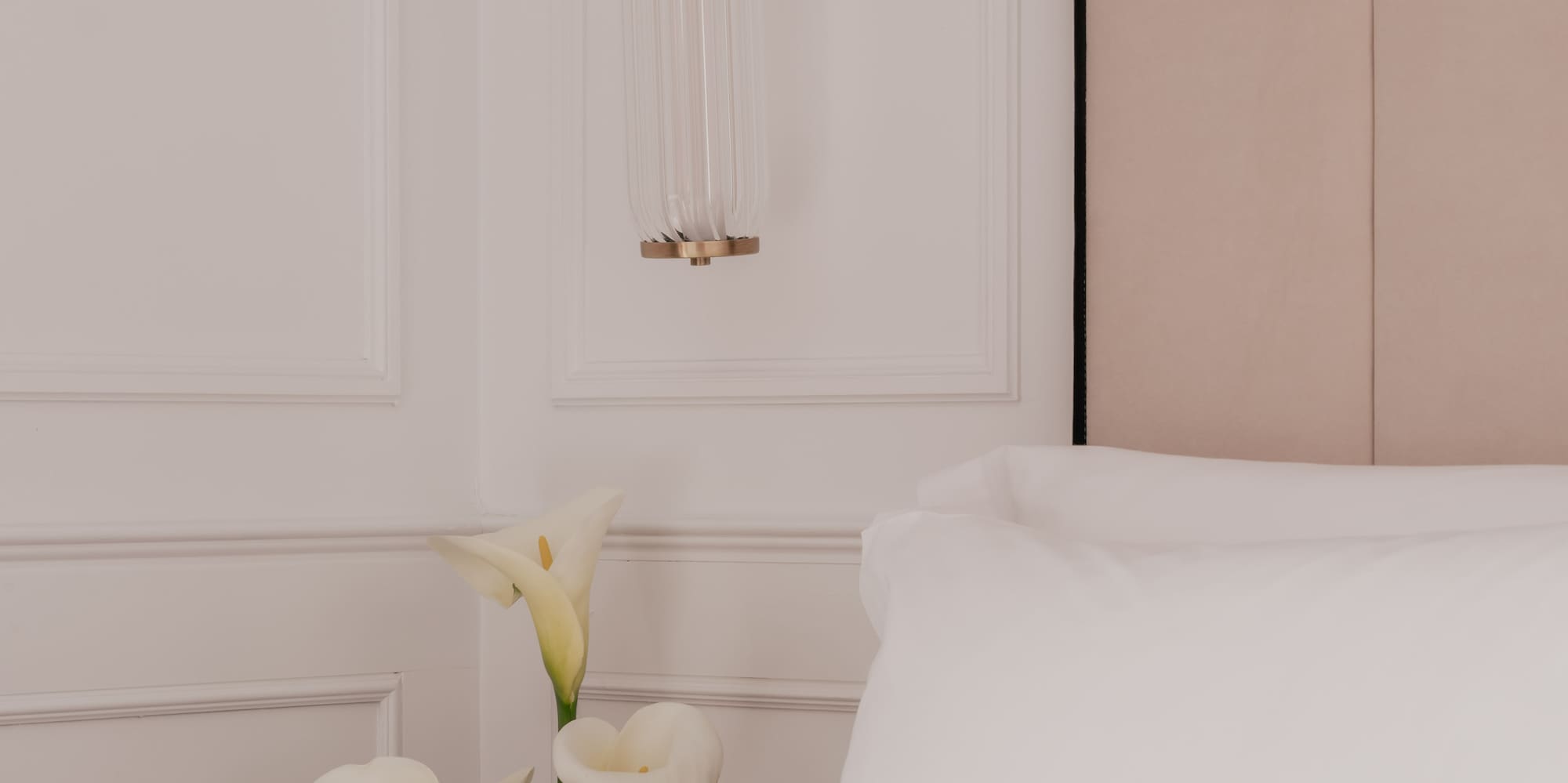 Modern bedroom with brass sconce light, calla lilies, white bedding.