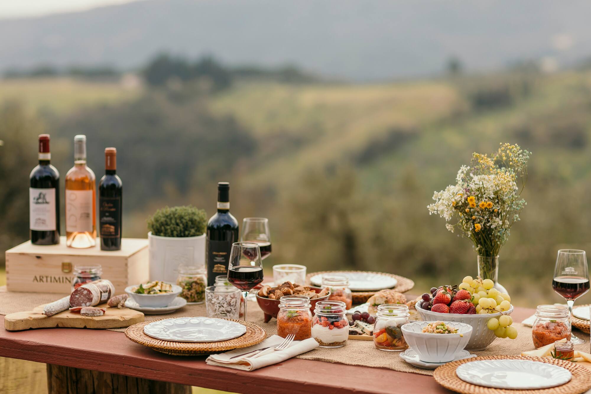 Rustic outdoor picnic table with wine, charcuterie, fruit, and snacks.
