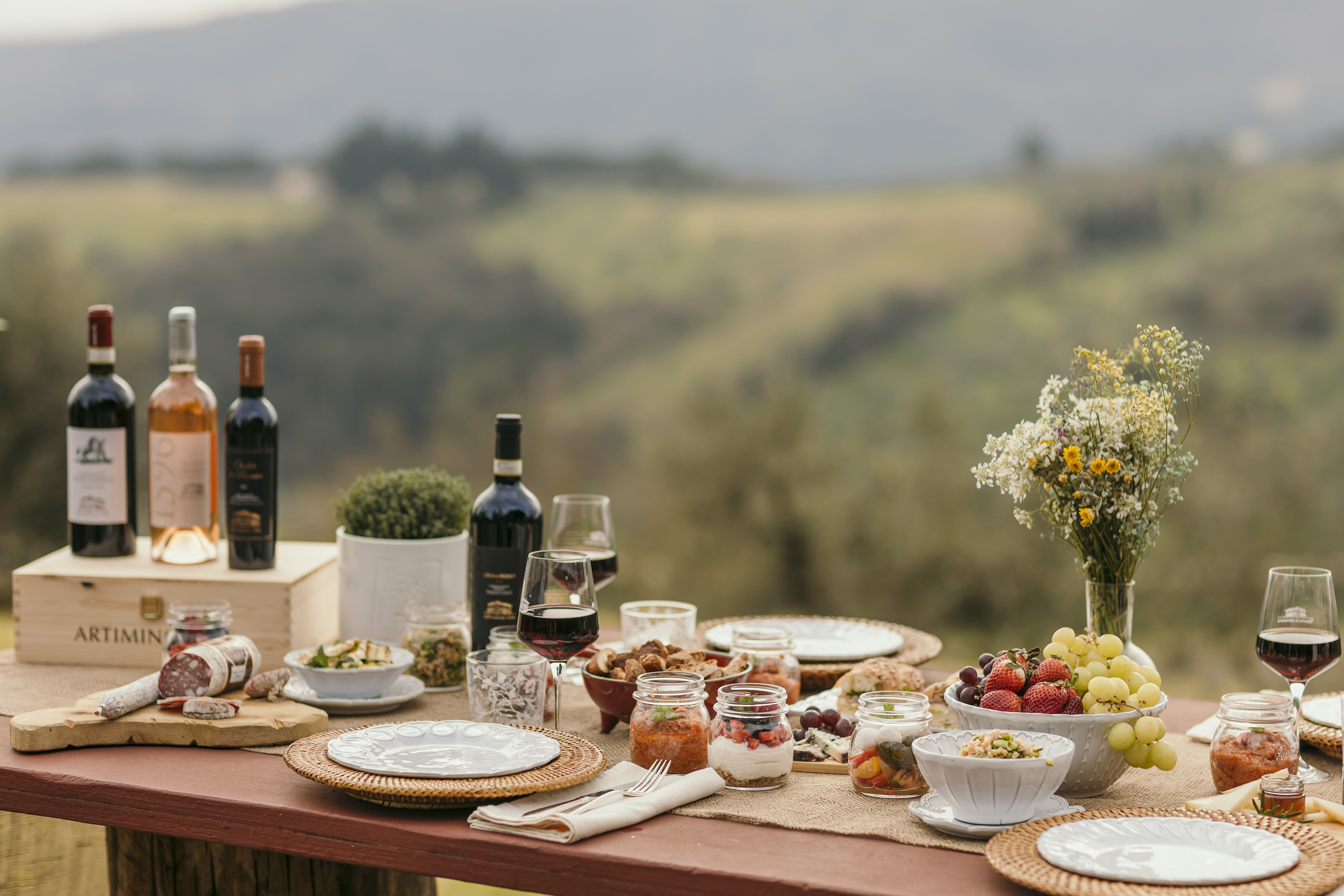 Rustic outdoor picnic table with wine, charcuterie, fruit, and snacks.