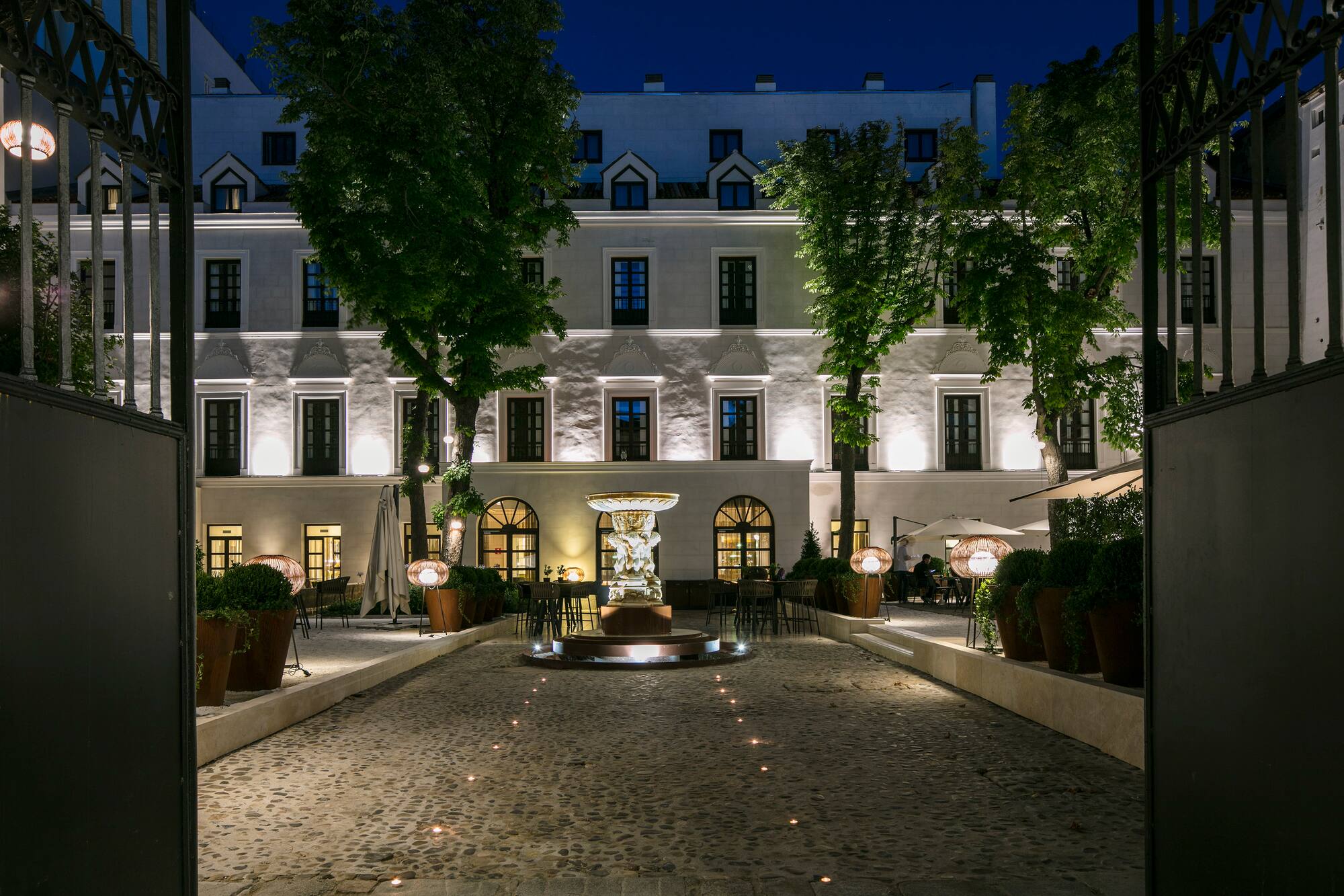 Hotel courtyard at night, illuminated fountain, cobblestone path, outdoor dining areas.