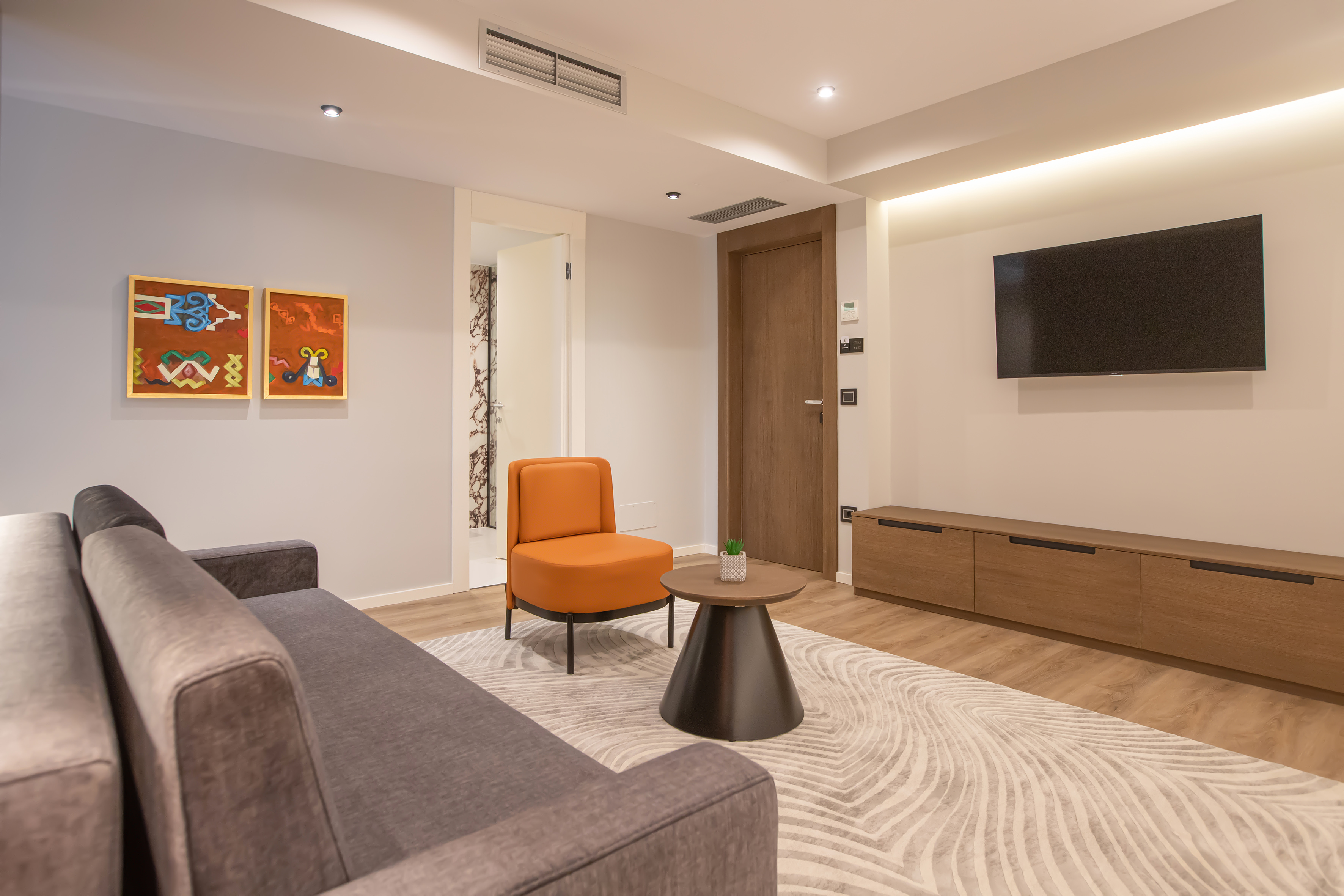 Modern apartment living room with grey sofa, orange chair, TV, and abstract art.