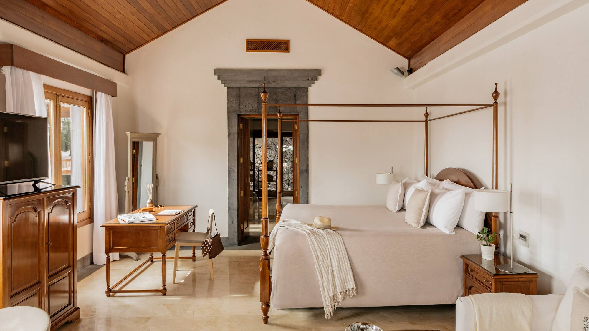 Luxury hotel bedroom with four-poster bed, vaulted wood ceiling, and stone doorway.