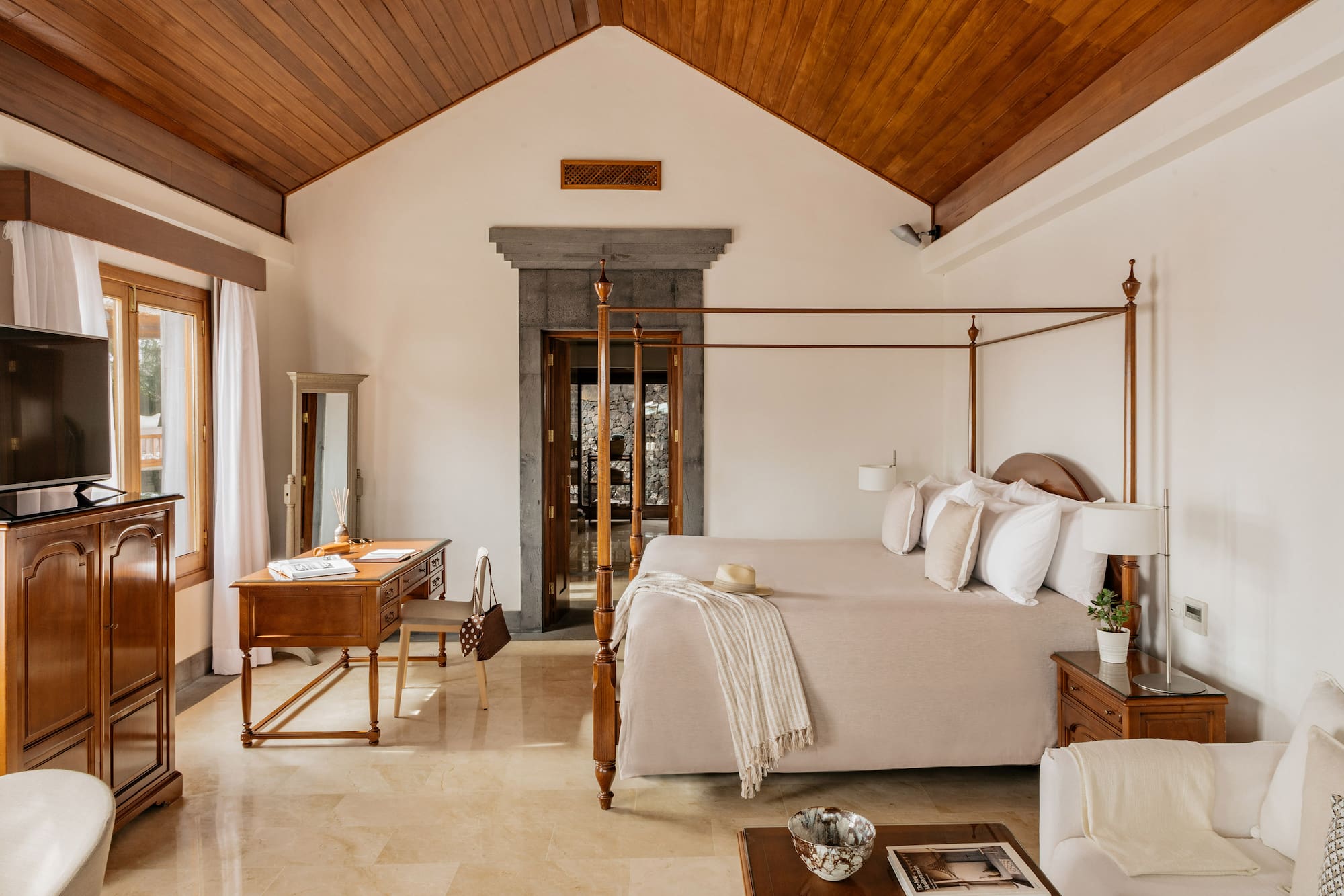 Luxury hotel bedroom with four-poster bed, vaulted wood ceiling, and stone doorway.