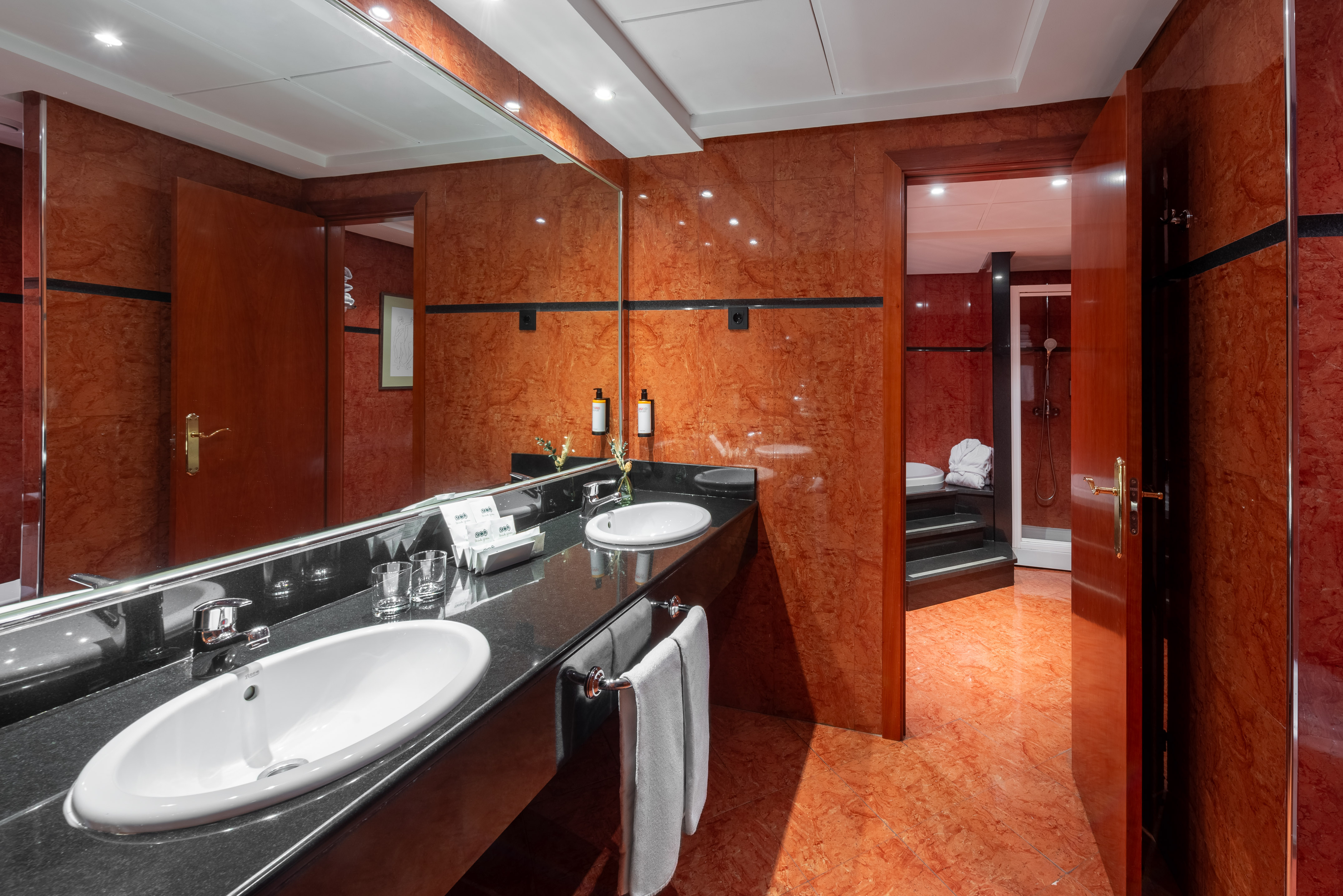 Modern hotel bathroom with red marble walls, double vanity, mirror, and shower area.