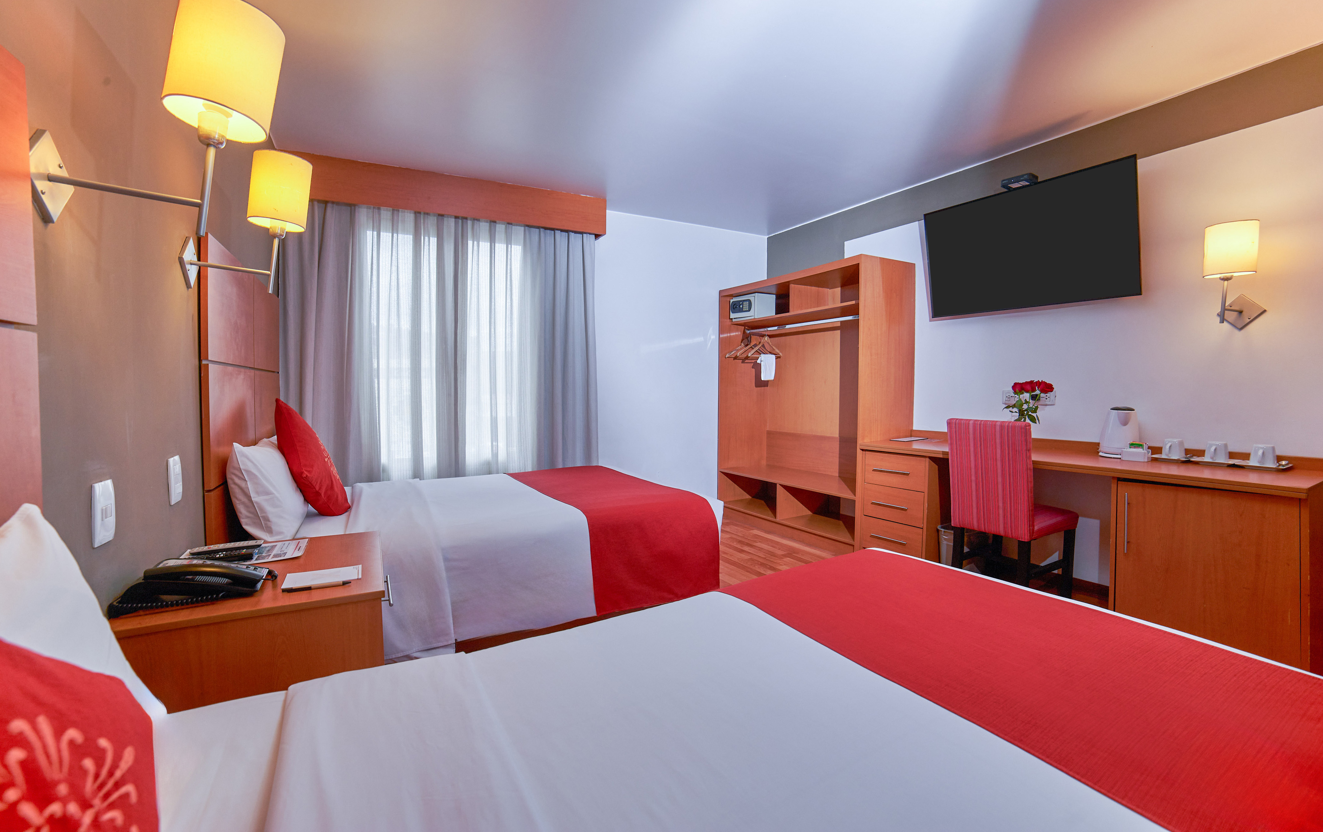 Comfortable twin hotel room with red accents, wall TV, desk, and wardrobe.