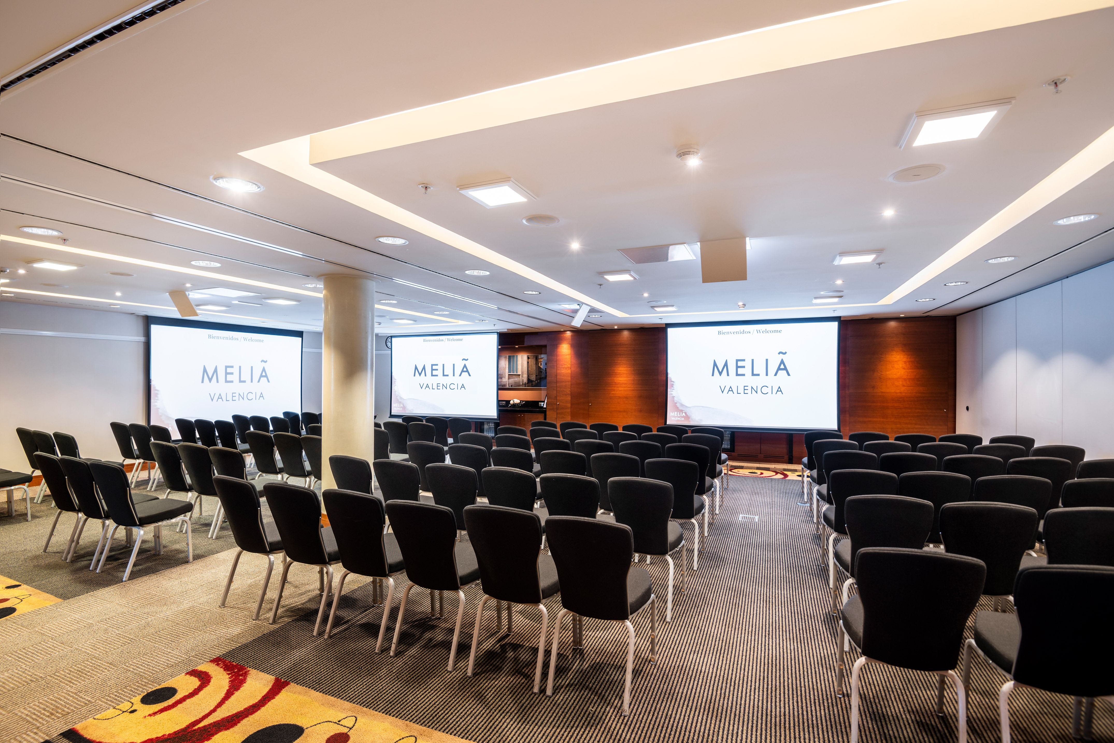 Empty Meliã Valencia hotel conference room, rows of chairs facing three screens.