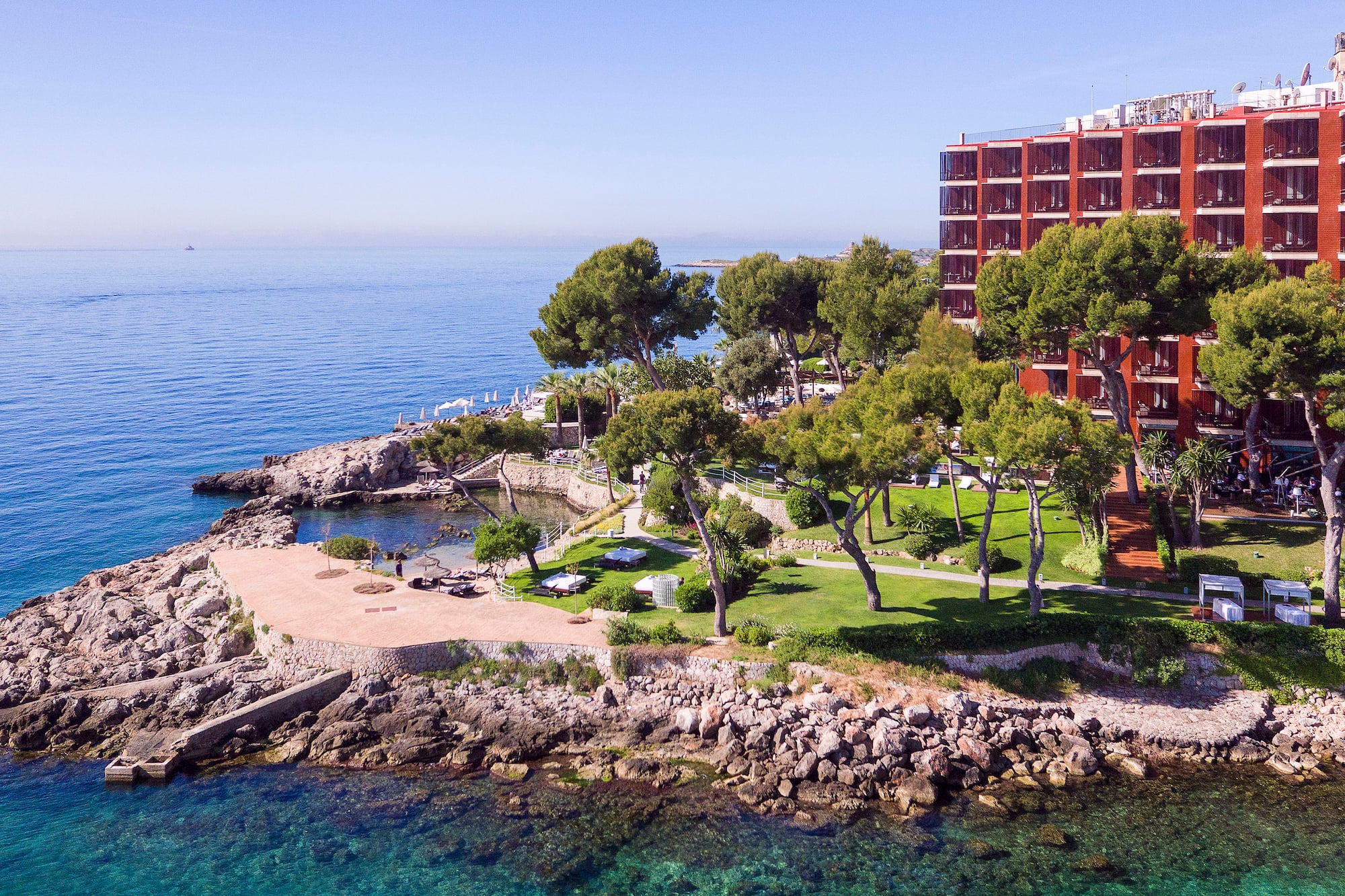 Red hotel on rocky coast with green trees and clear blue ocean.