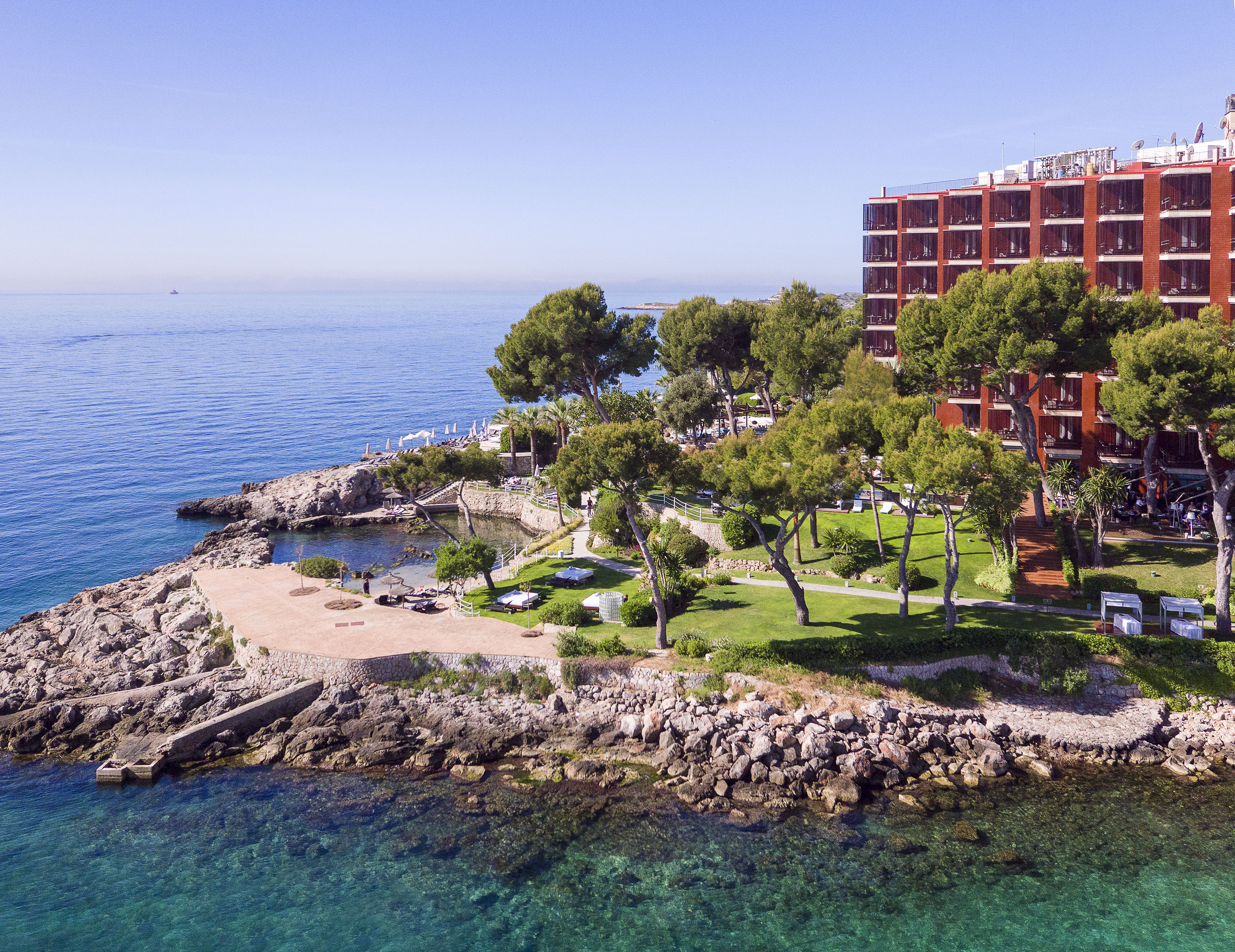 Red hotel on rocky coast with green trees and clear blue ocean.