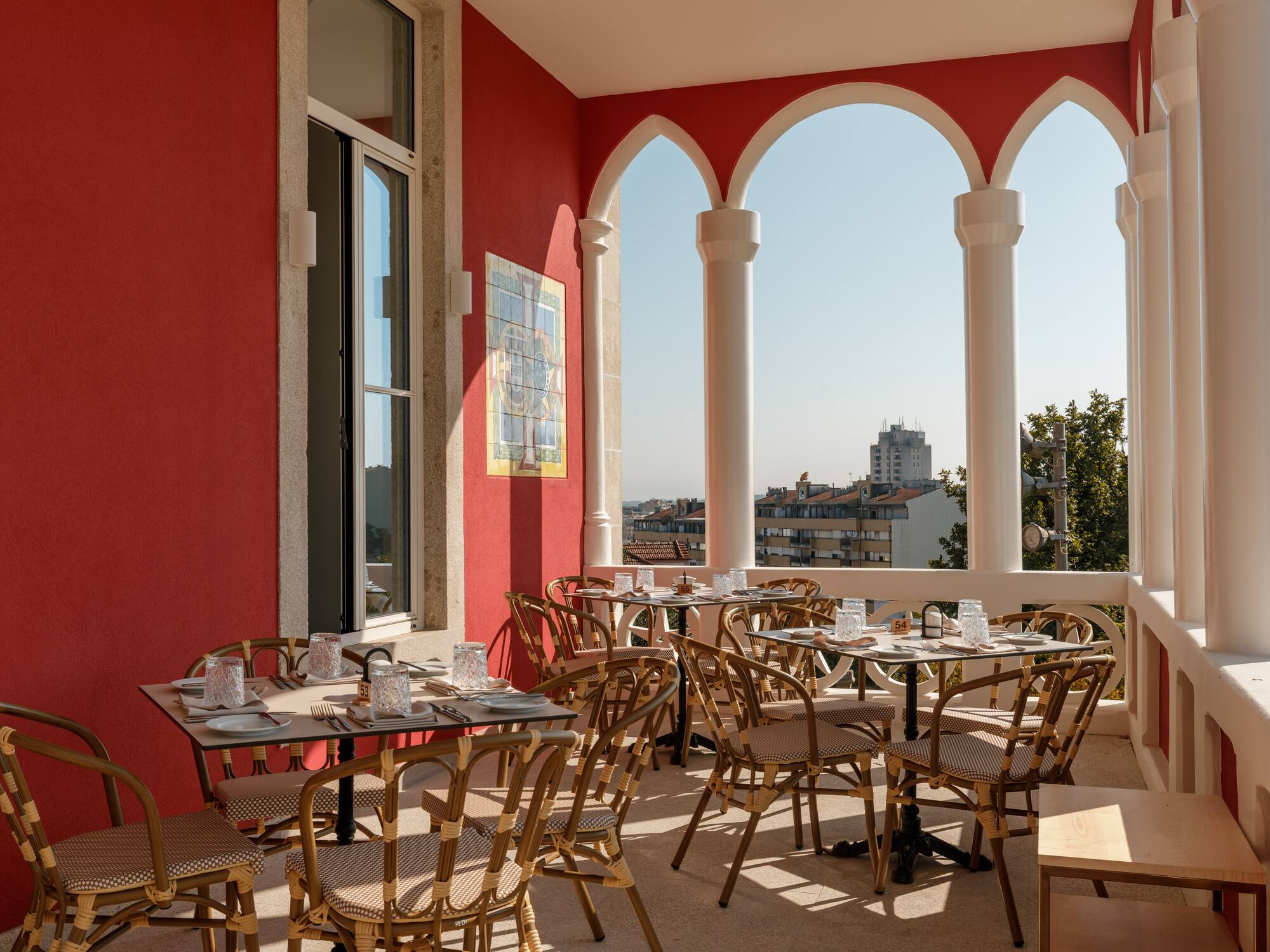 Red-walled outdoor dining terrace with arched columns, rattan chairs, and city view.