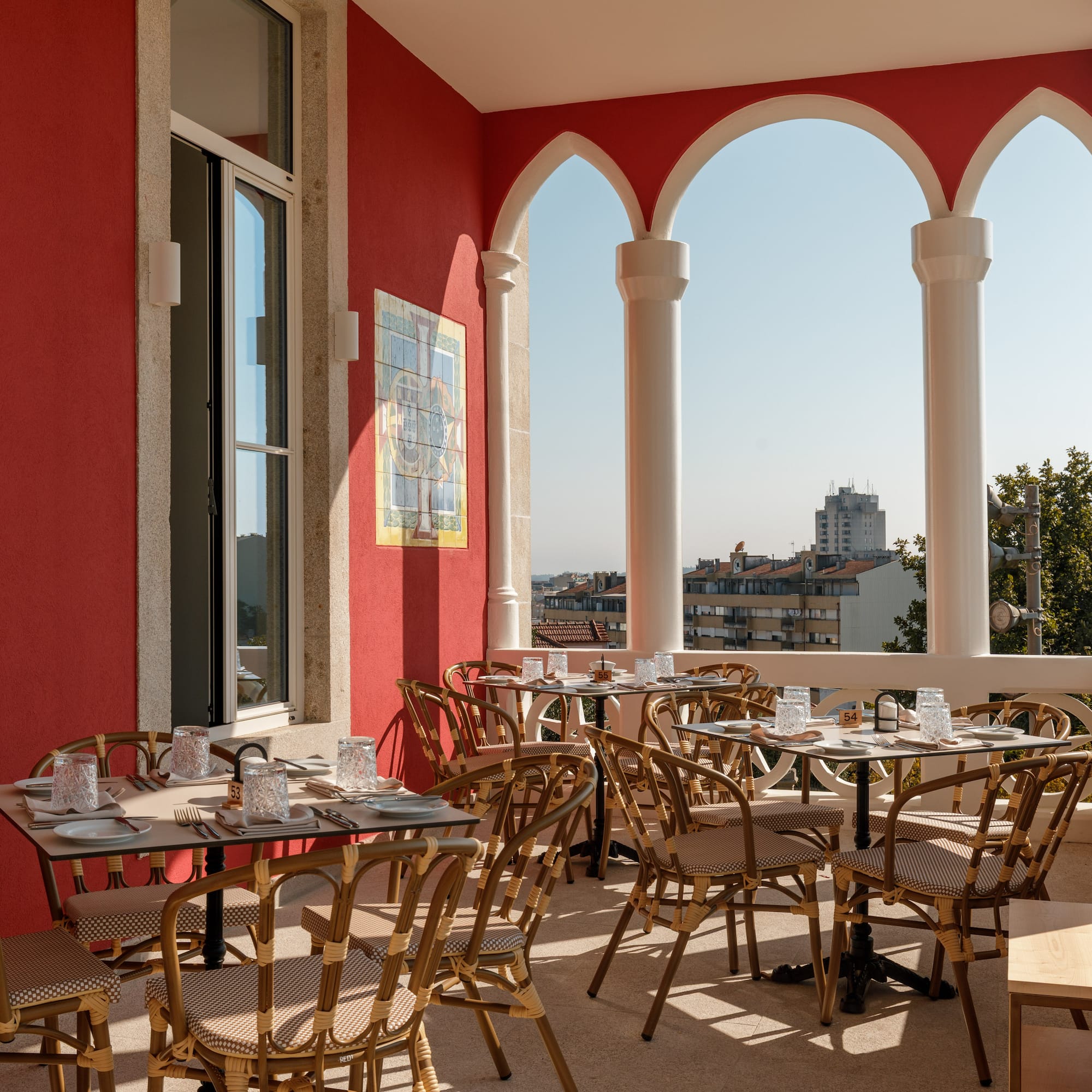 Red-walled outdoor dining terrace with arched columns, rattan chairs, and city view.