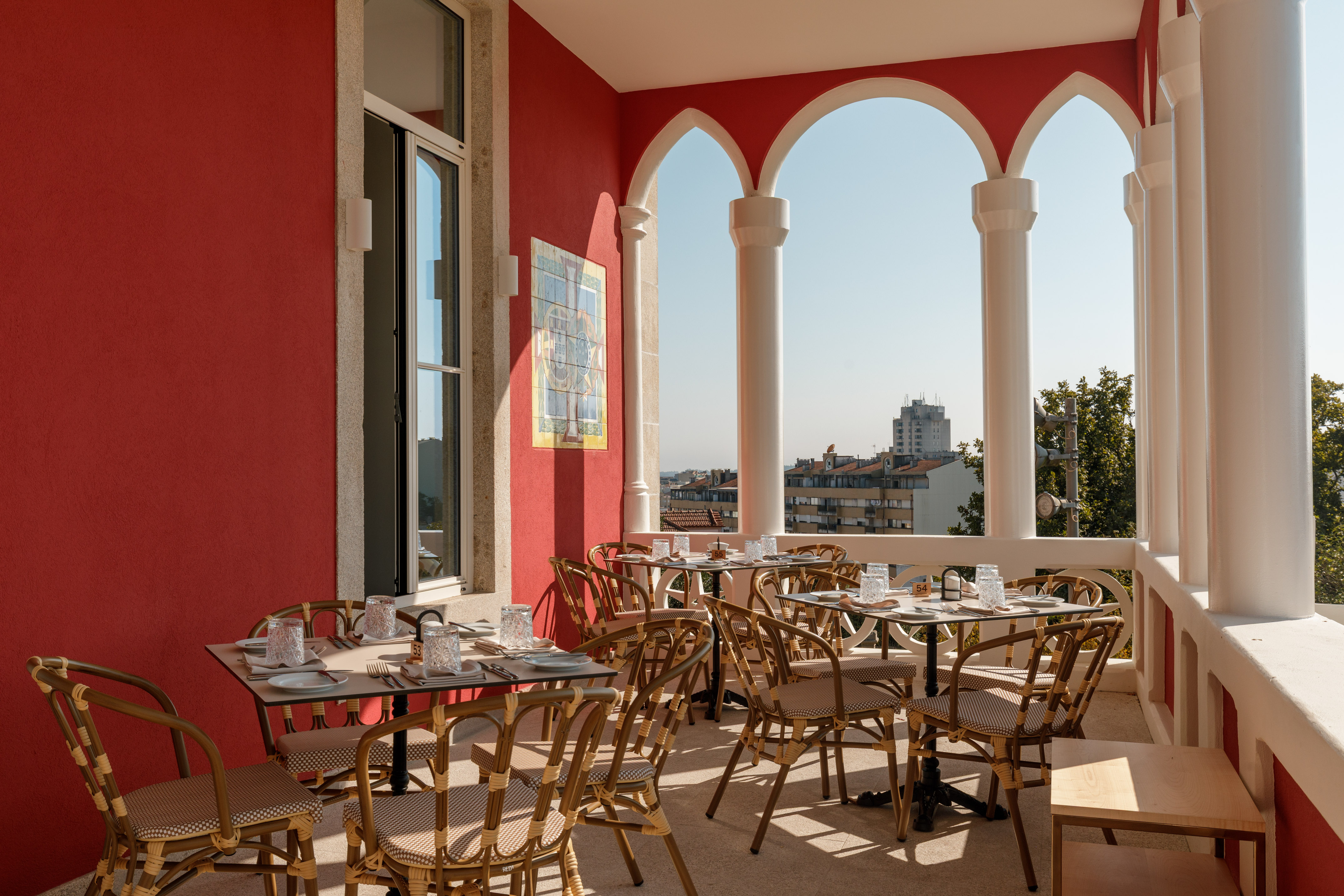 Red-walled outdoor dining terrace with arched columns, rattan chairs, and city view.