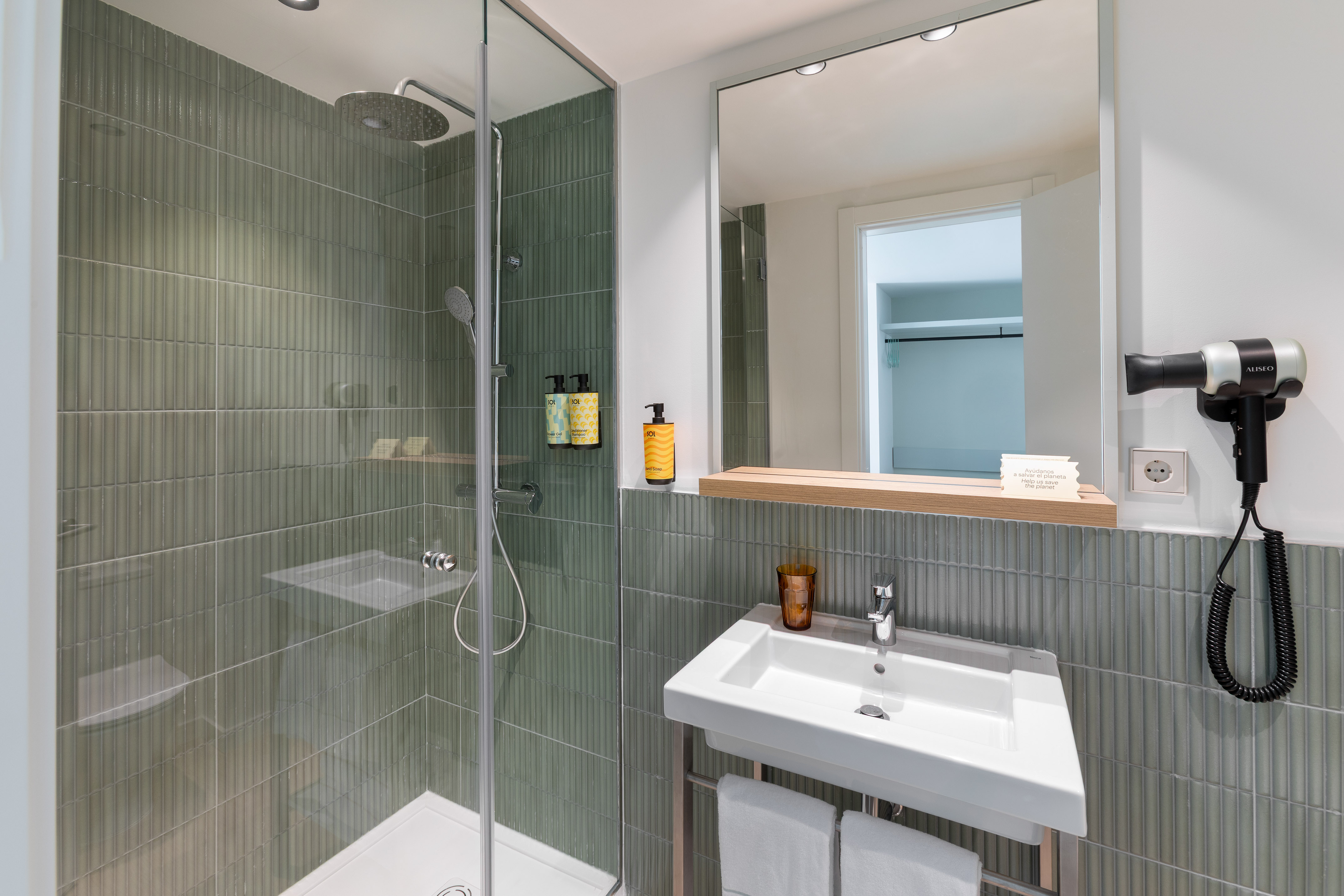 Modern hotel bathroom with green tiled shower, white vanity sink, and mirror.