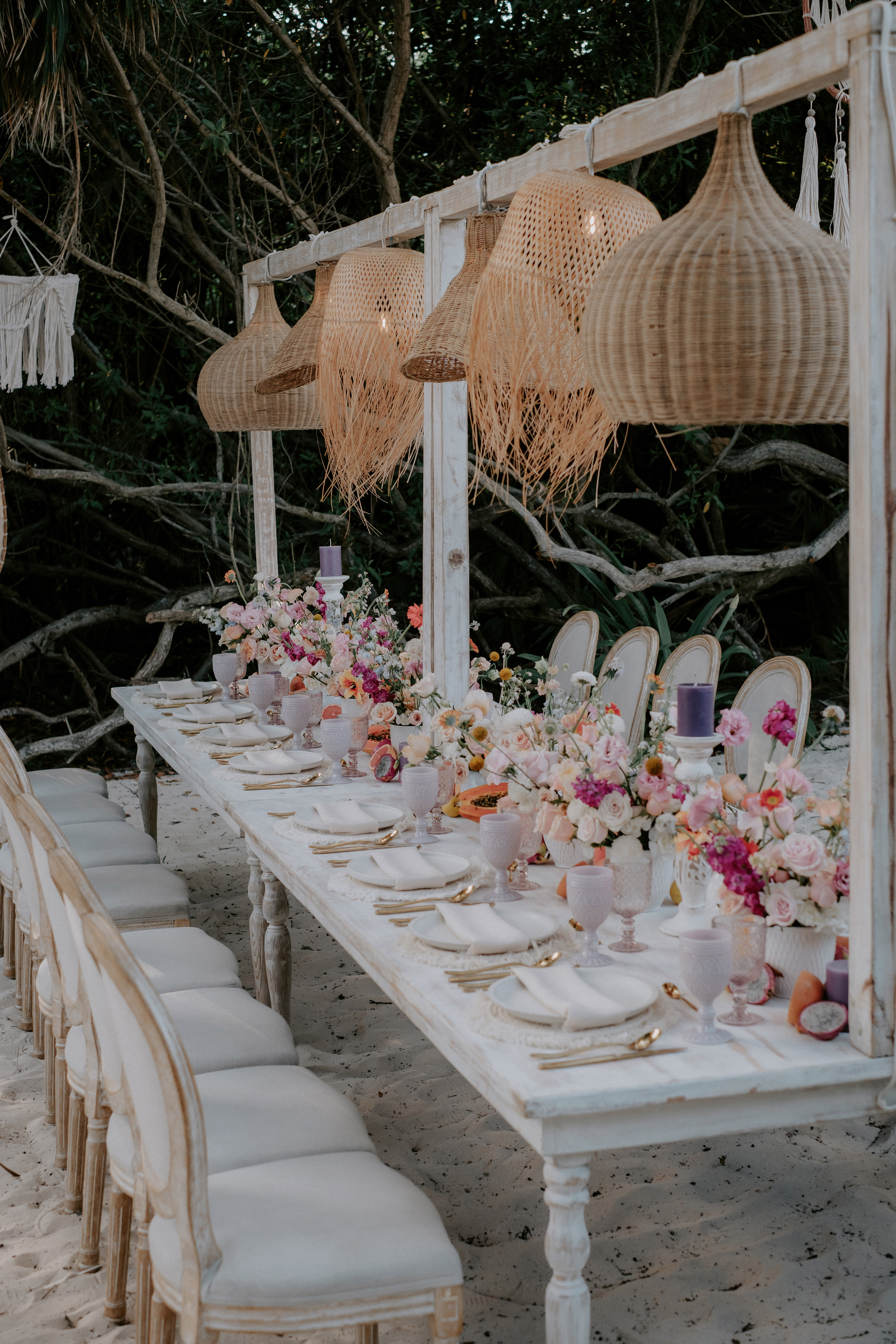 Beach wedding dinner table, white chairs, floral centerpieces, rattan lights.