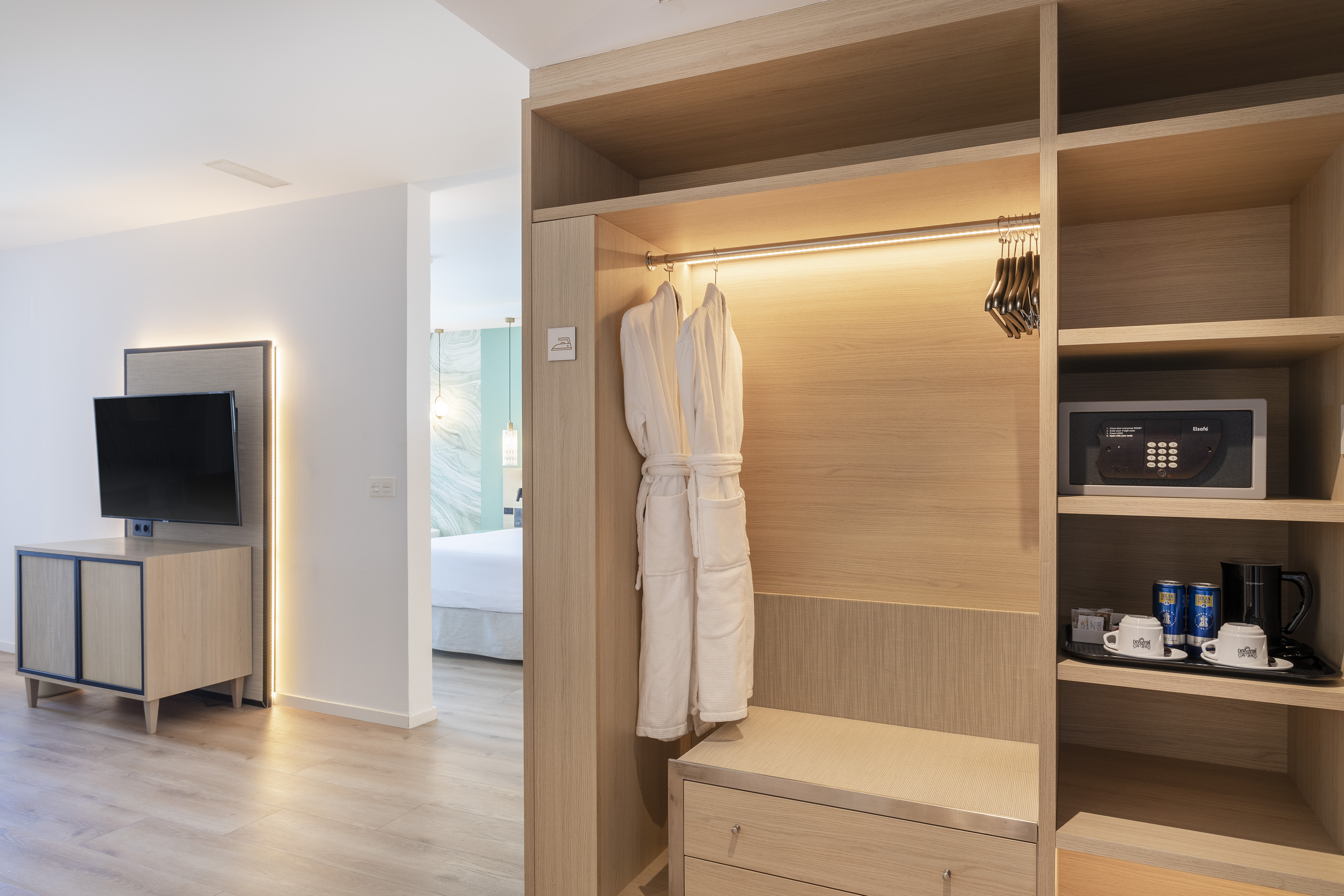Modern hotel room closet with bathrobes, safe, minibar, and TV.