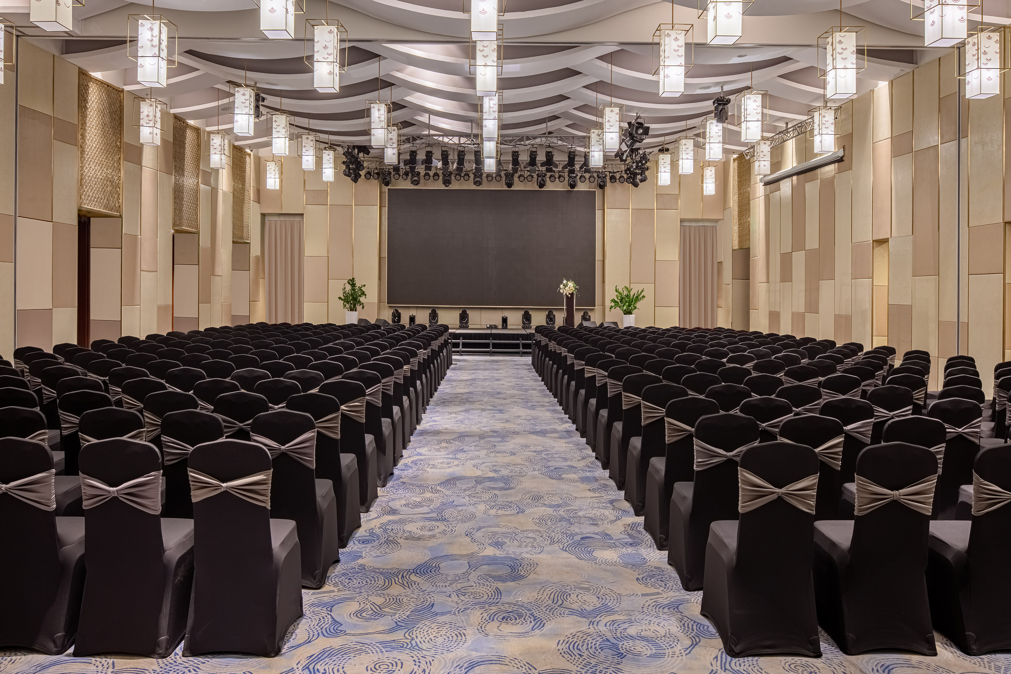 Empty conference hall with rows of black chairs, stage, and large display screen.