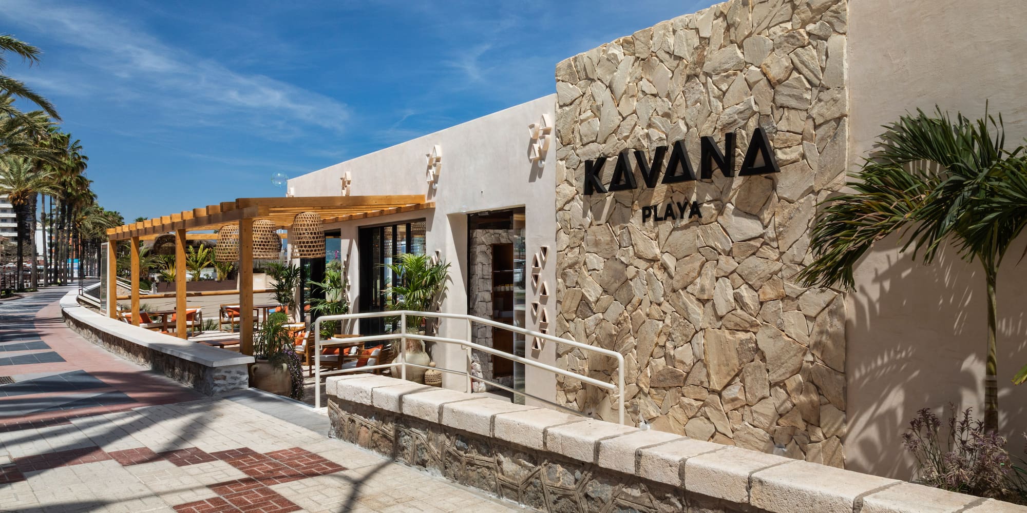 Kavana Playa restaurant exterior with stone wall, outdoor pergola, and palm trees.