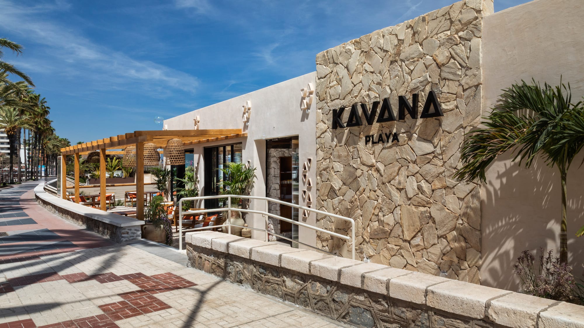 Kavana Playa restaurant exterior with stone wall, outdoor pergola, and palm trees.