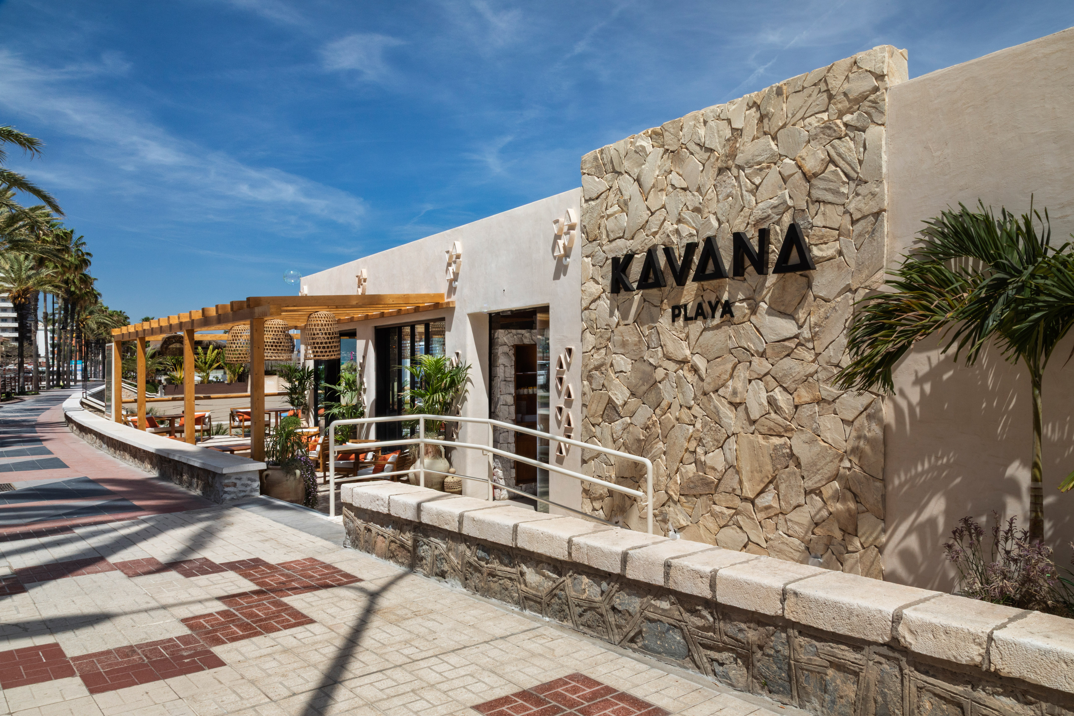 Kavana Playa restaurant exterior with stone wall, outdoor pergola, and palm trees.