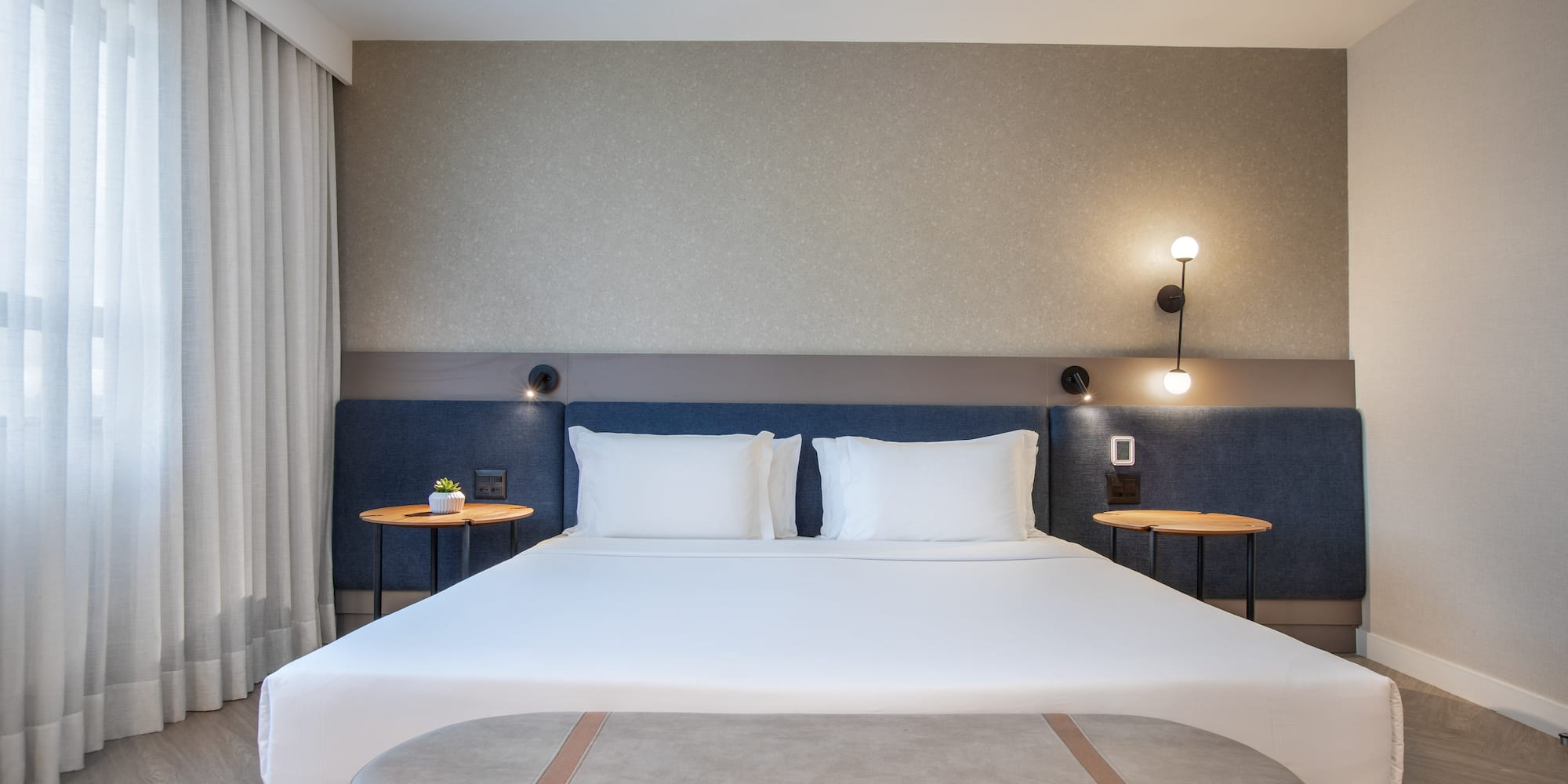 Modern hotel bedroom with large bed, blue headboard, nightstands, and bench.