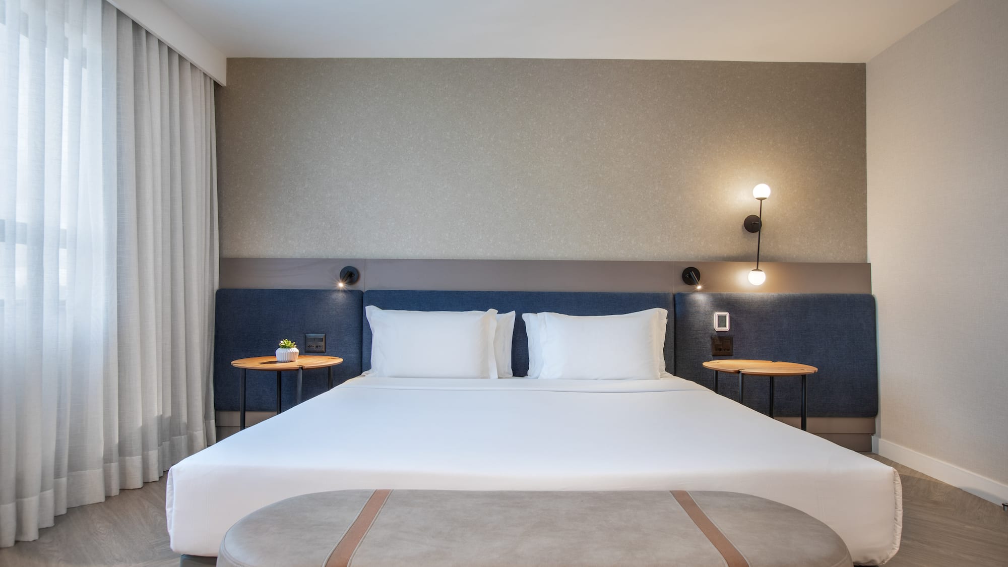 Modern hotel bedroom with large bed, blue headboard, nightstands, and bench.