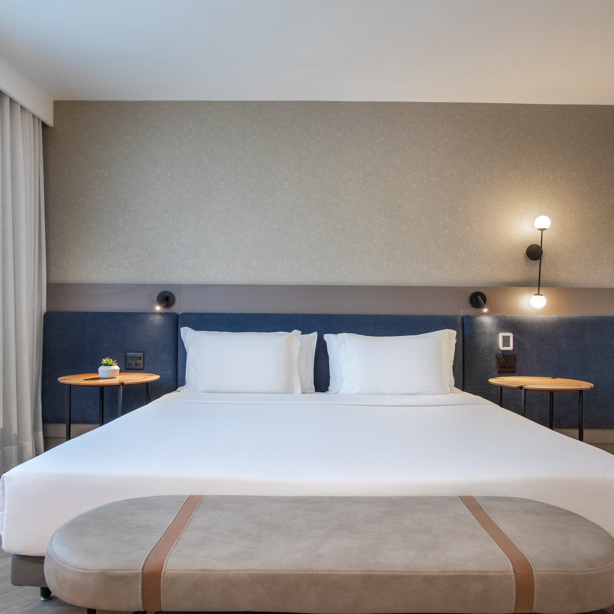 Modern hotel bedroom with large bed, blue headboard, nightstands, and bench.