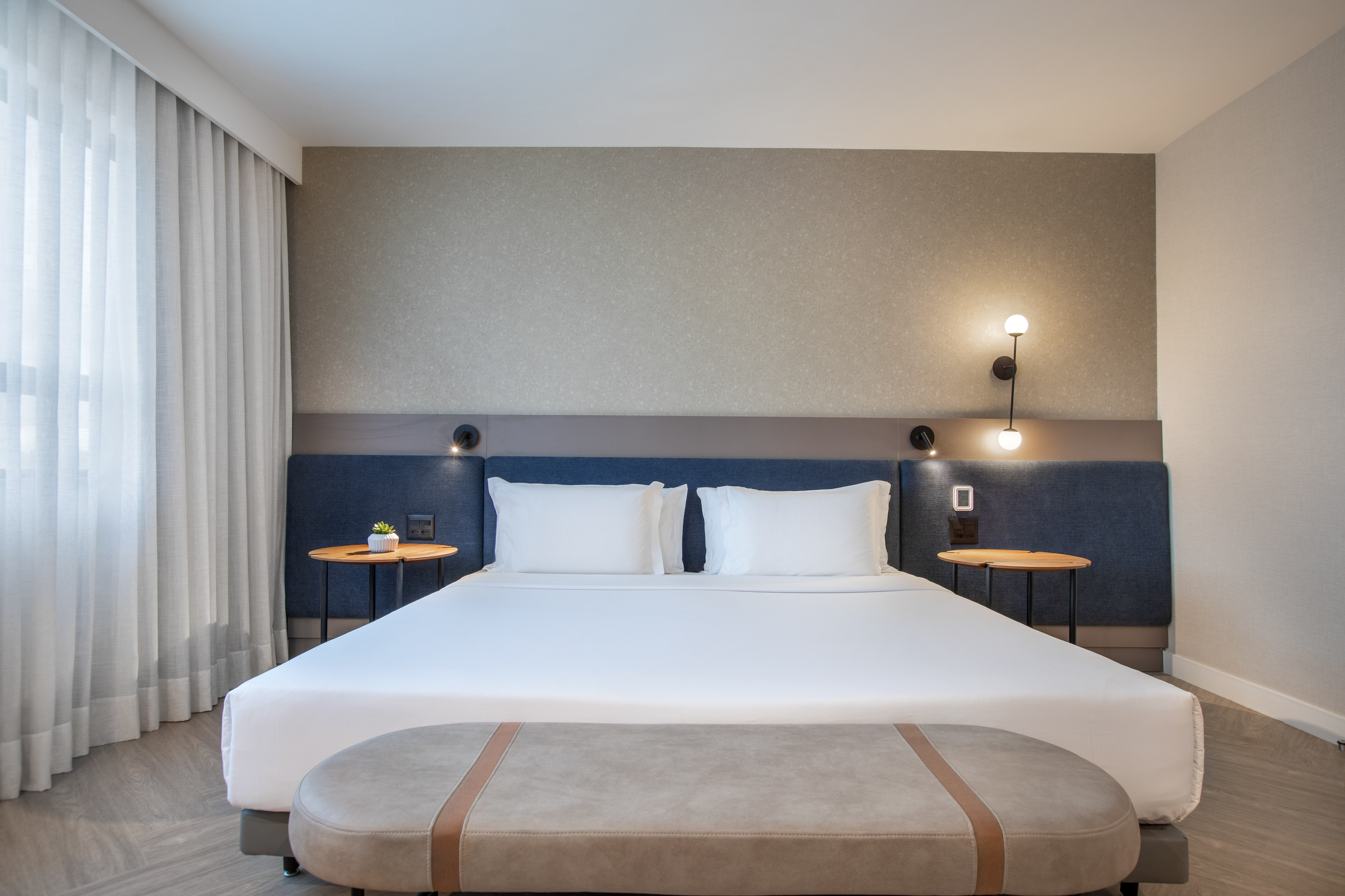 Modern hotel bedroom with large bed, blue headboard, nightstands, and bench.
