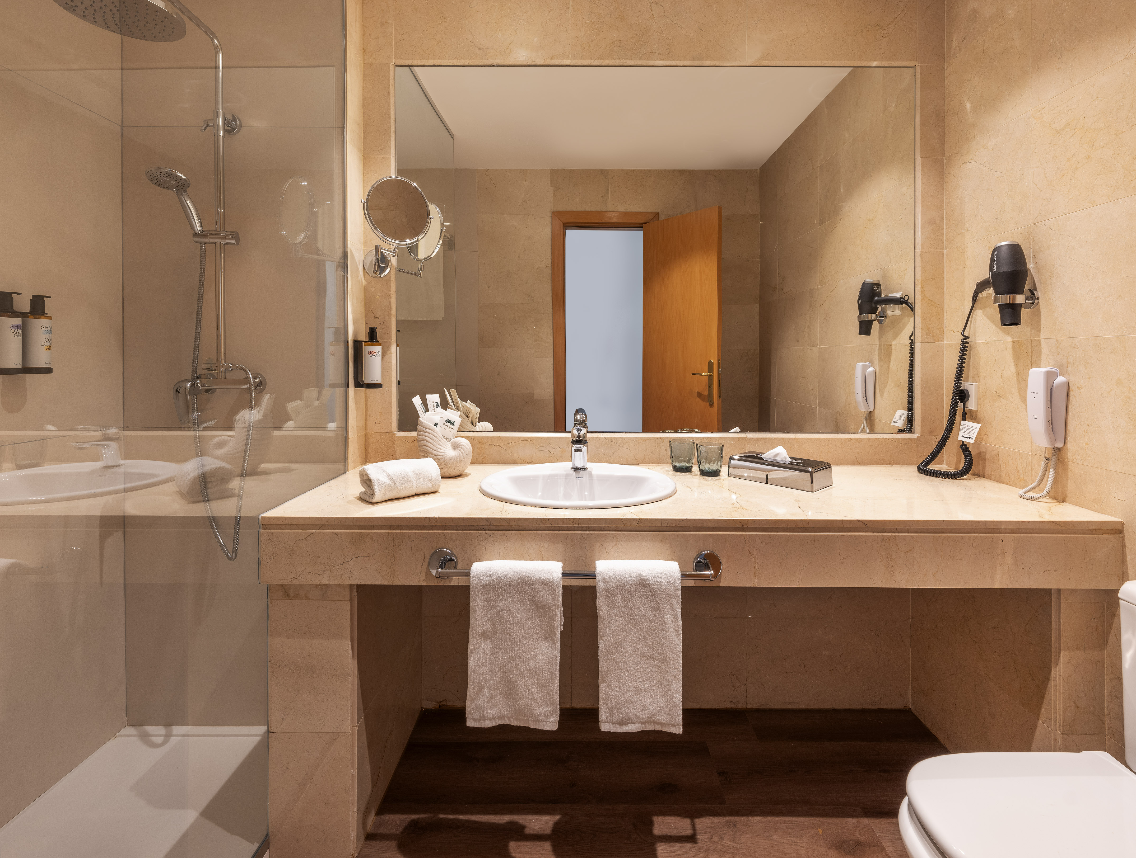 Modern hotel bathroom with glass shower, vanity, large mirror, and amenities.