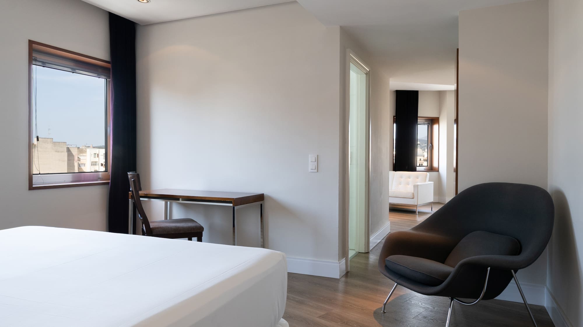 Modern hotel room with bed, desk, city view window, and lounge armchair.