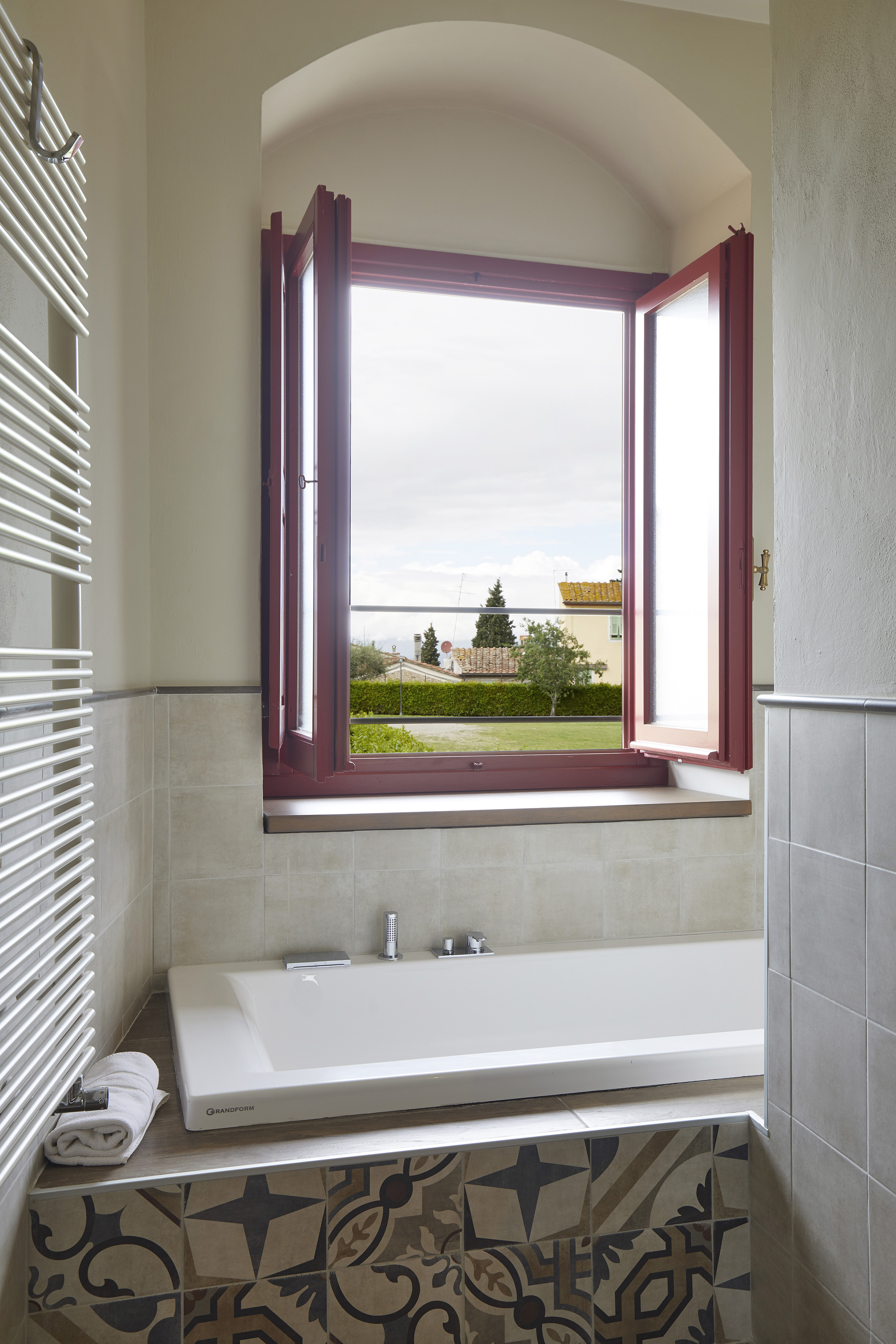 Bathroom with white bathtub, open red-framed window to countryside, patterned tiles.