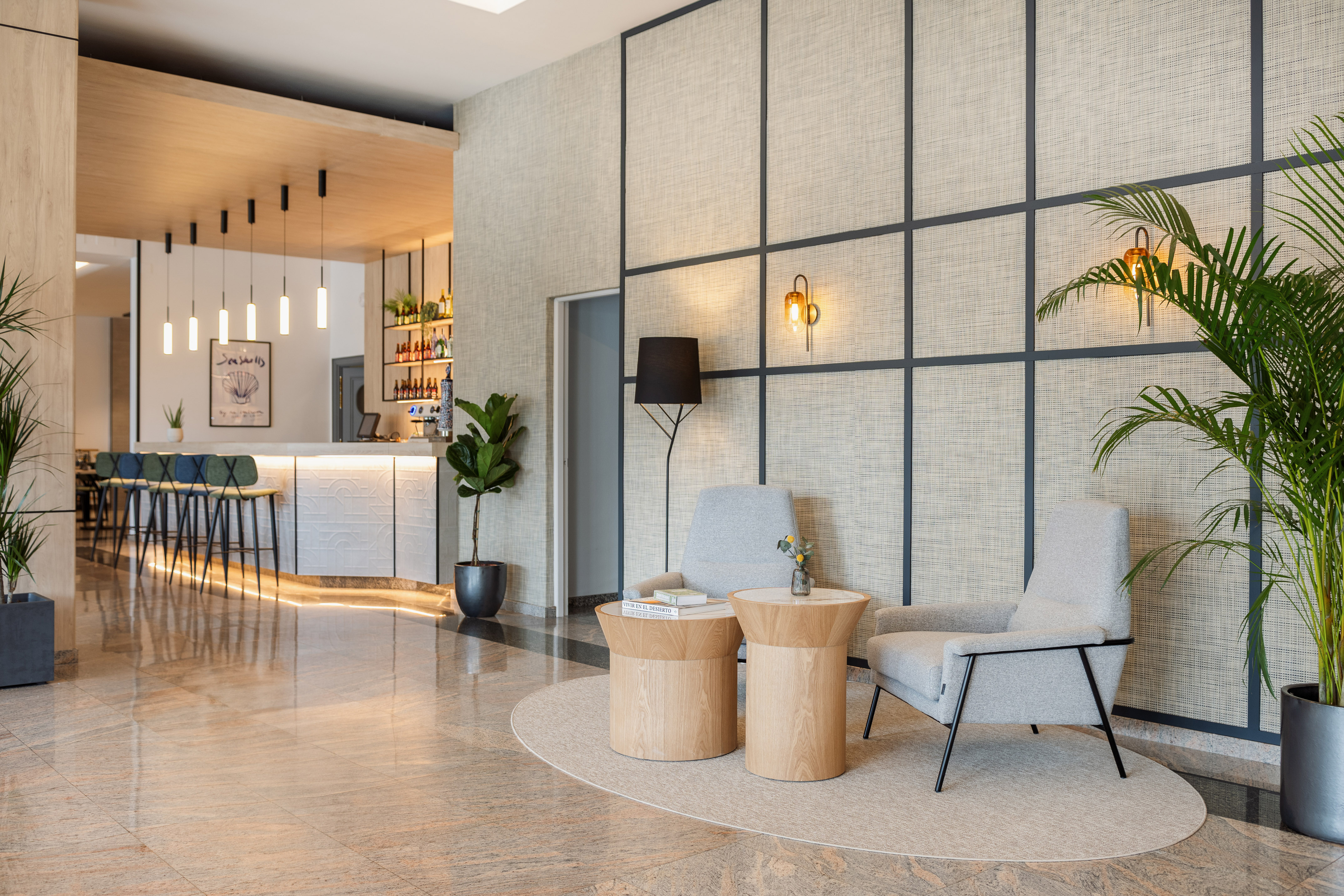 Modern hotel lobby lounge with bar, armchairs, wooden tables, and plants.