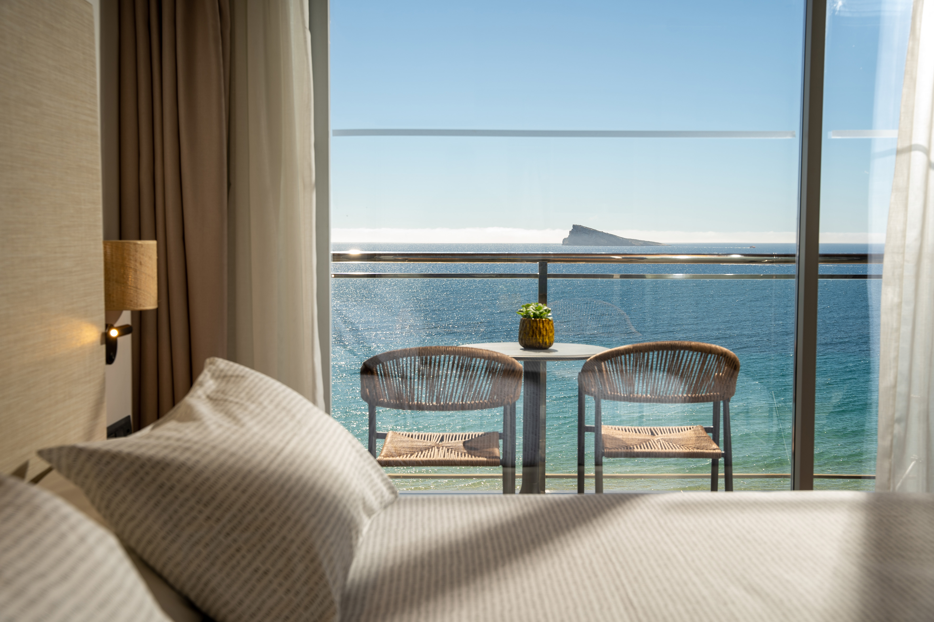 Hotel room balcony with ocean view, chairs, and distant island.