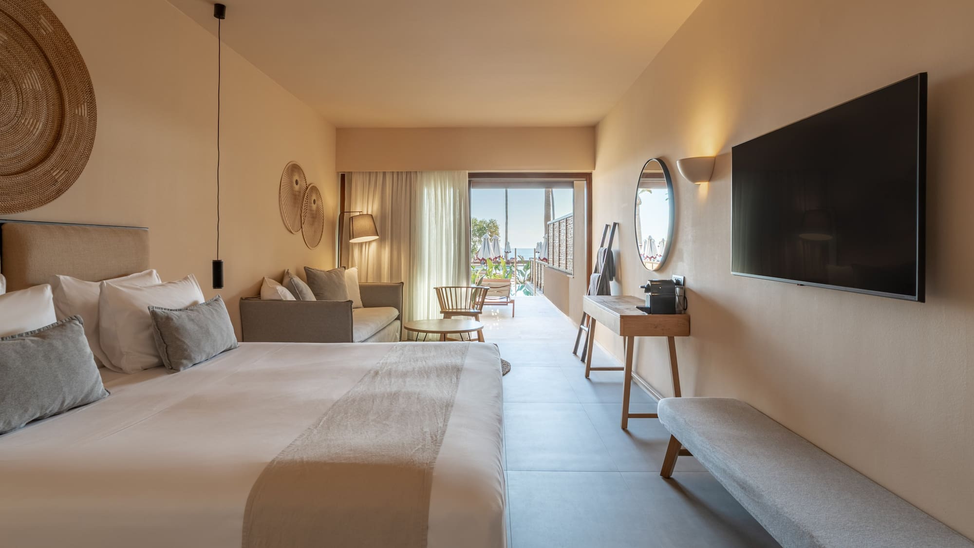 Hotel suite with modern bedroom, sofa, TV, and ocean view balcony.