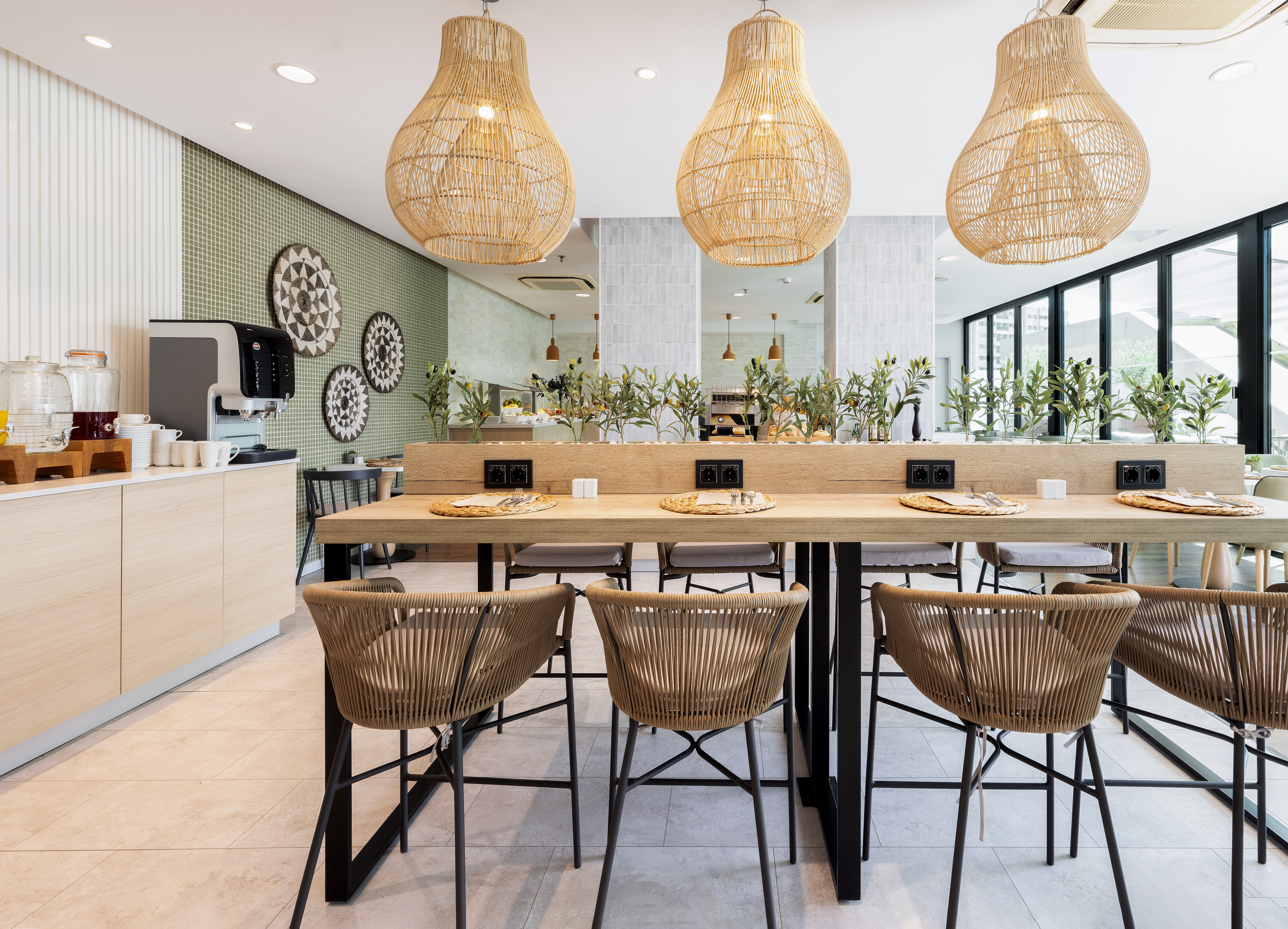 Modern hotel breakfast bar with long table, rattan chairs, and woven lights.
