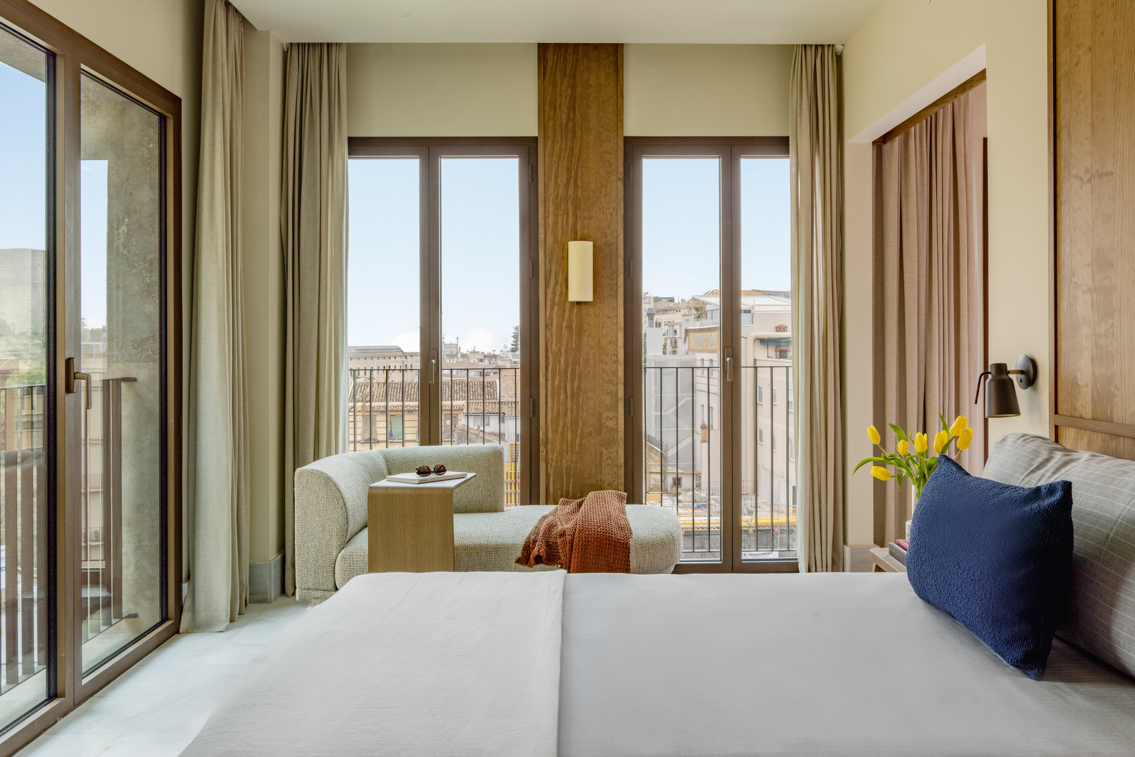 Modern hotel bedroom with city view balcony, chaise lounge, and bed.