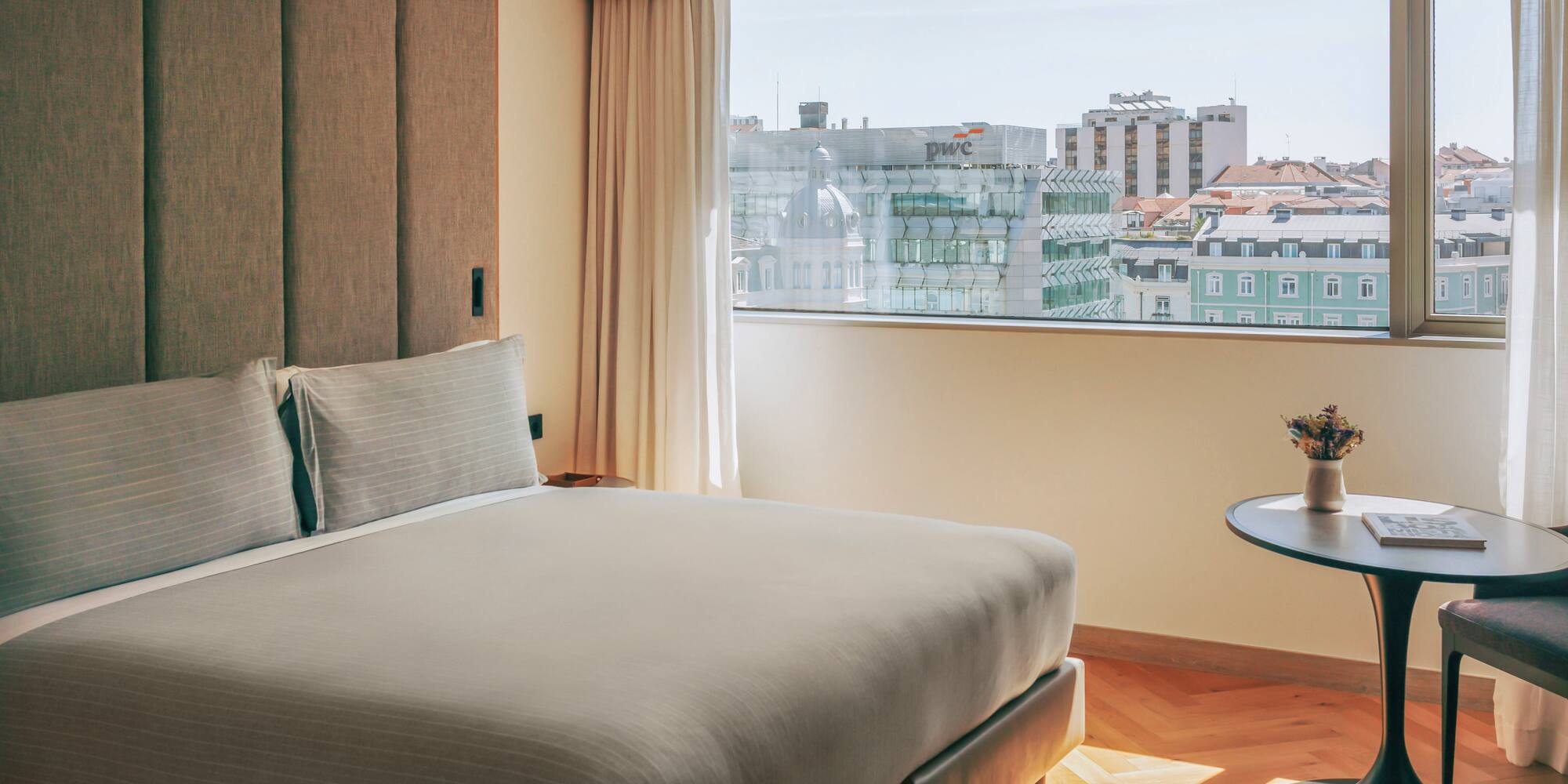 Hotel bedroom with modern bed, textured headboard, and a bright city view.