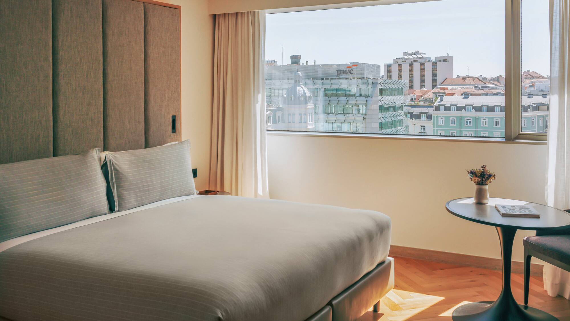 Hotel bedroom with modern bed, textured headboard, and a bright city view.
