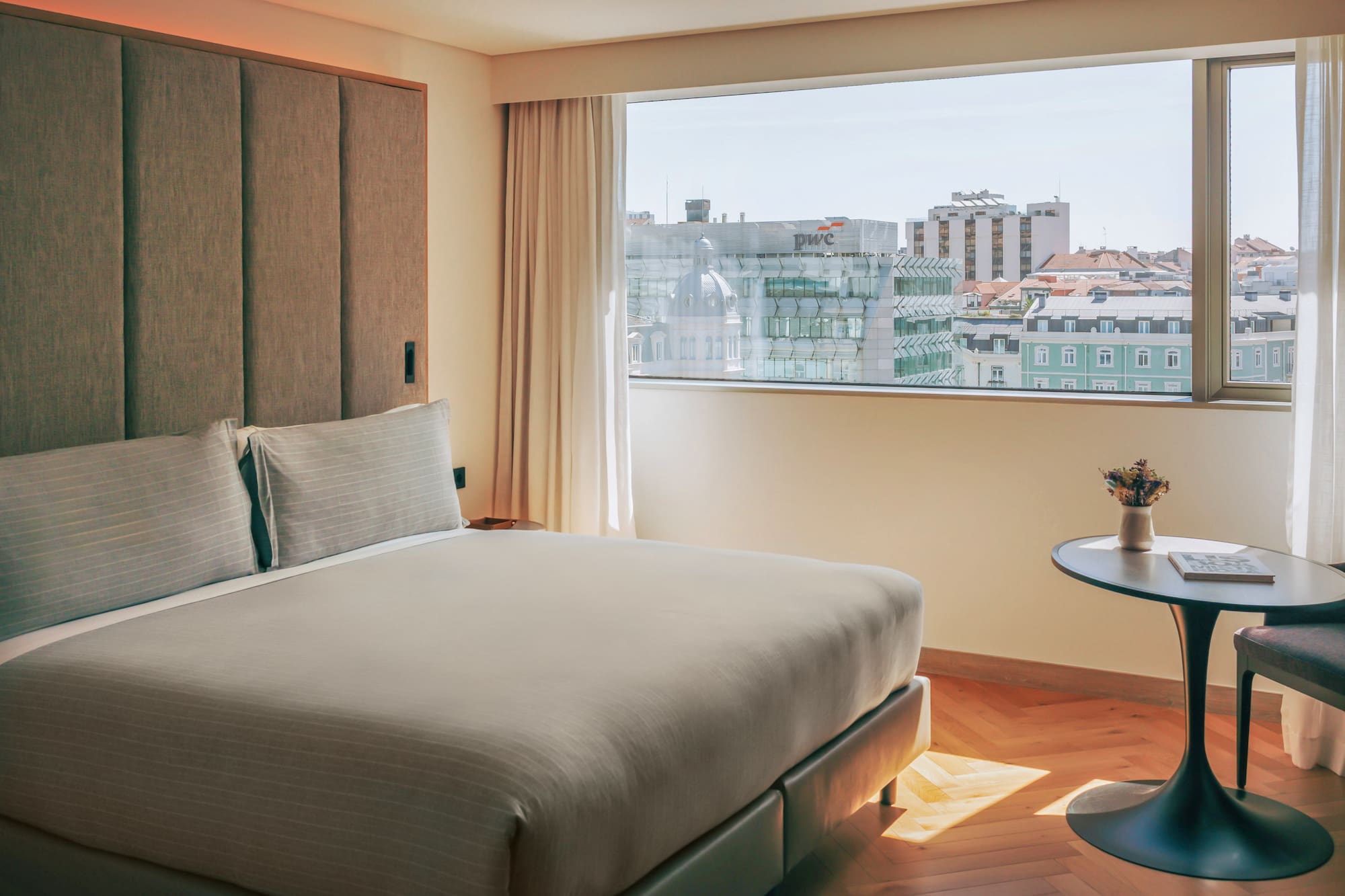 Hotel bedroom with modern bed, textured headboard, and a bright city view.