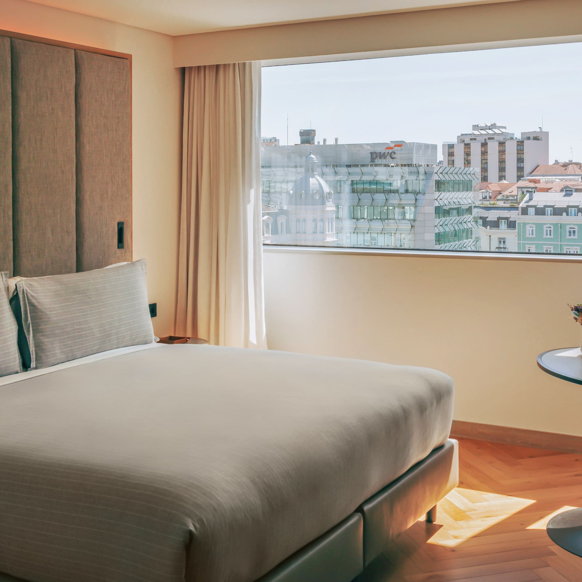 Hotel bedroom with modern bed, textured headboard, and a bright city view.