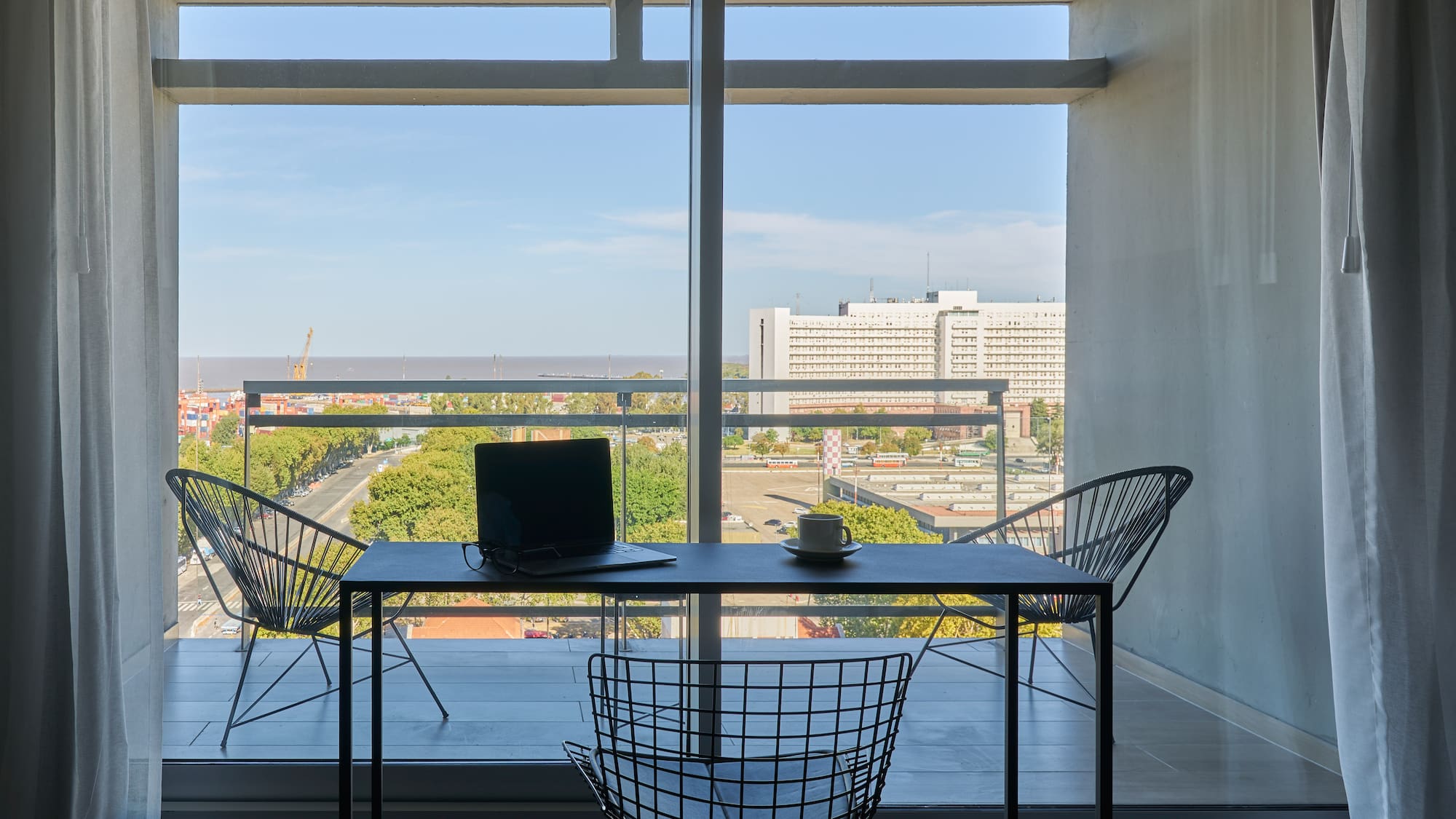 Balcony home office with laptop, coffee, and urban water view.